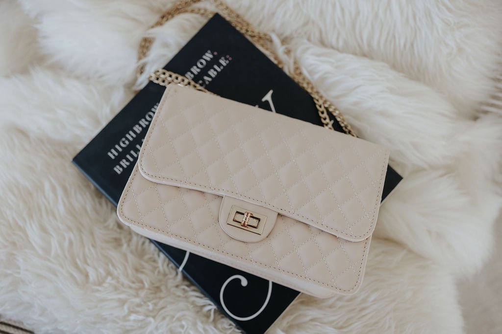 White quilted clutch purse with a gold clasp resting on a black book with white and pink text, on a fluffy white surface.