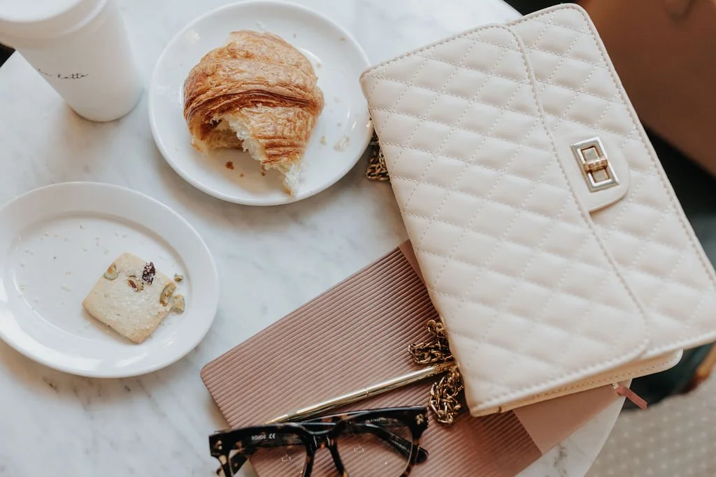 A white marble table with a coffee cup, a plate with a croissant, a plate with a partially eaten snack, a beige quilted purse, a pink notebook, and a pair of glasses.