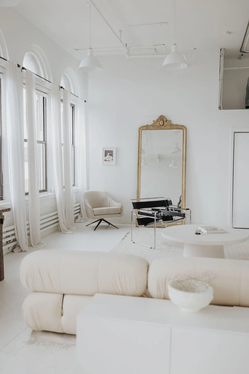 Bright, minimalist living room with white walls, high windows with white curtains, a large ornate gold mirror leaning against the wall, modern black and chrome chair, beige armchair, white couch, framed art, and a round white coffee table.
