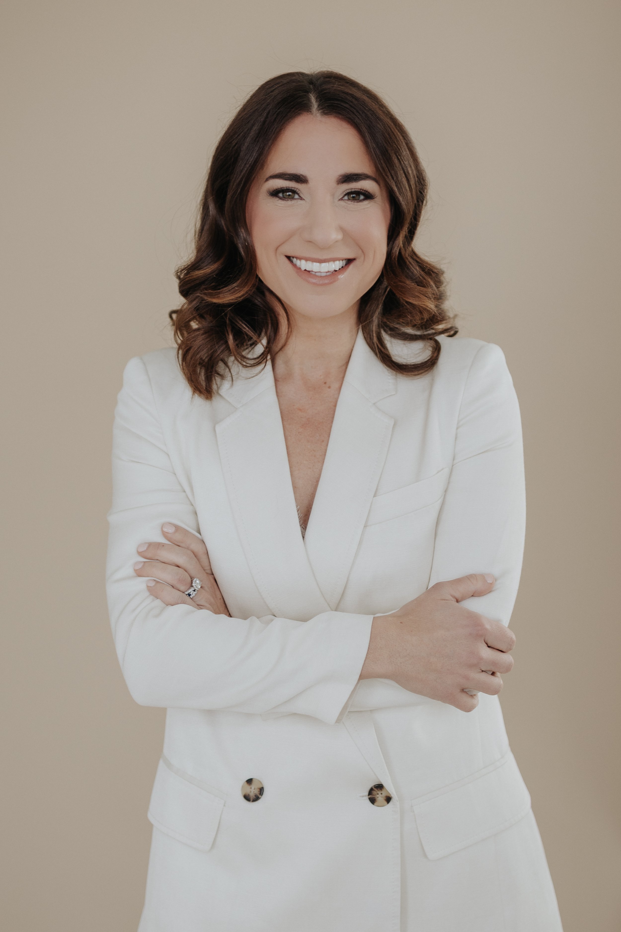 A woman with shoulder-length brown hair, wearing a white blazer and skirt, smiling with arms crossed against a beige background.