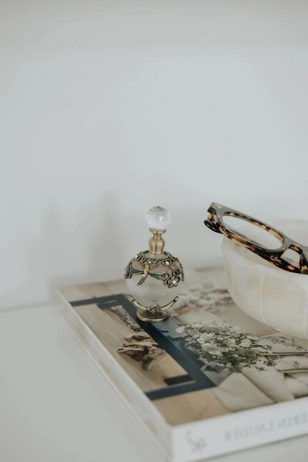 A decorative glass perfume bottle with a jeweled metal overlay and a glass stopper, a pair of tortoiseshell glasses, and a marble bowl resting on top of a magazine with various photographs and text, all placed on a white surface against a plain white wall.