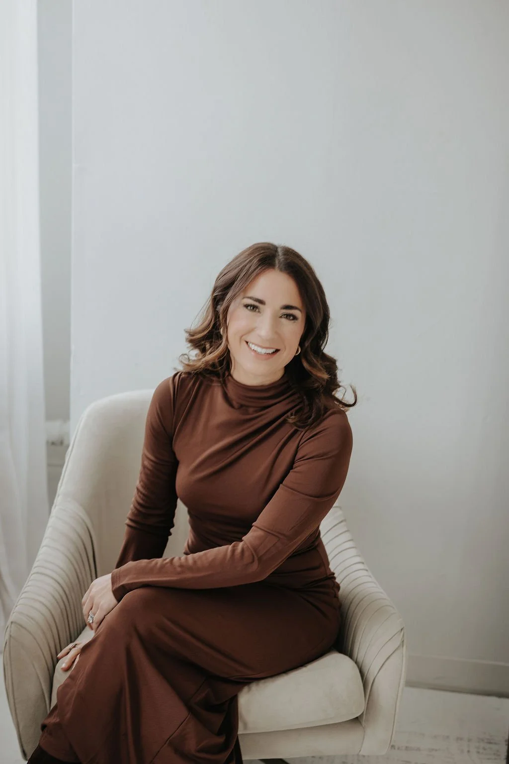 A woman with shoulder-length brunette hair and a warm smile, sitting on a white upholstered armchair in a minimalist room with white walls, wearing a chocolate brown long-sleeve dress.