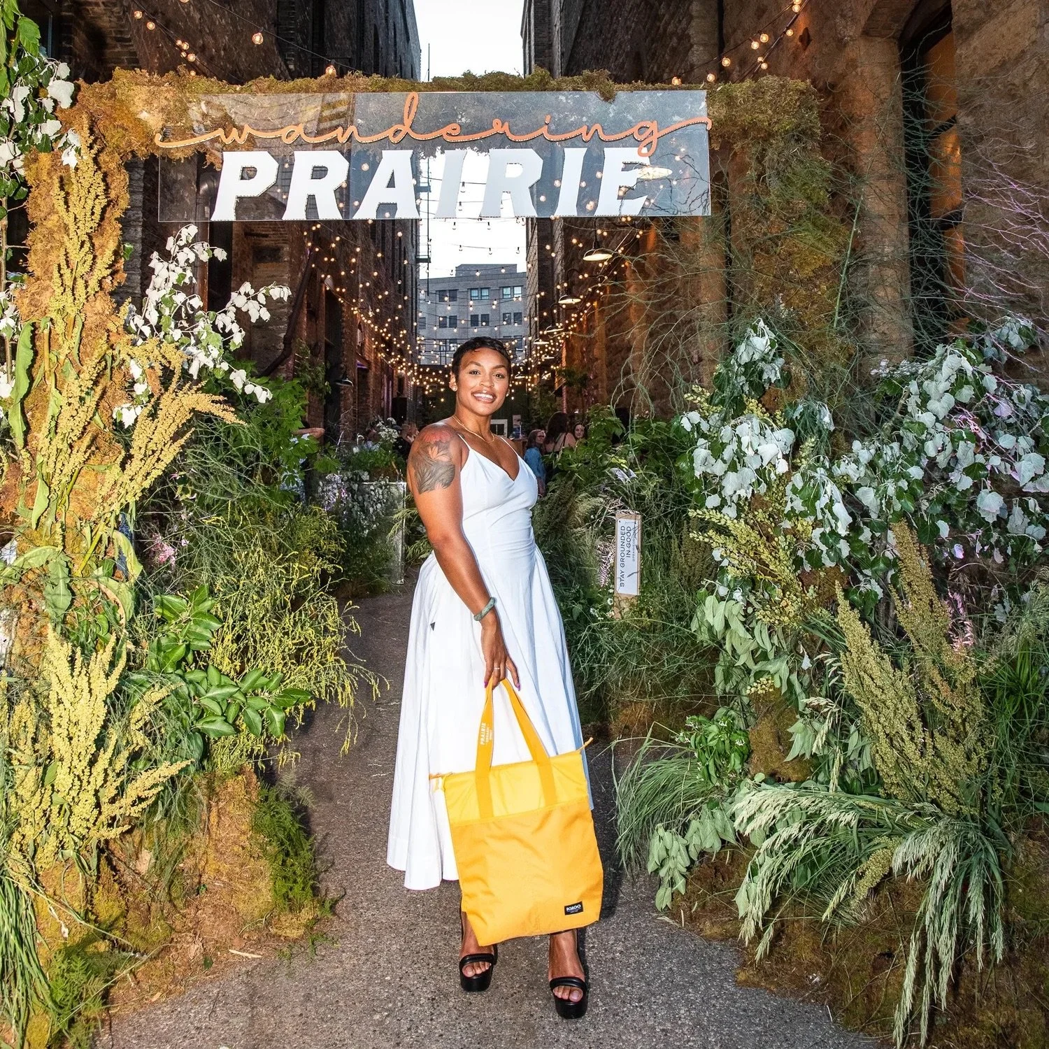 A woman in a white dress holding a yellow tote bag standing in a lush, decorated outdoor space with a sign that says "wandering PRAIRIE" and string lights overhead.