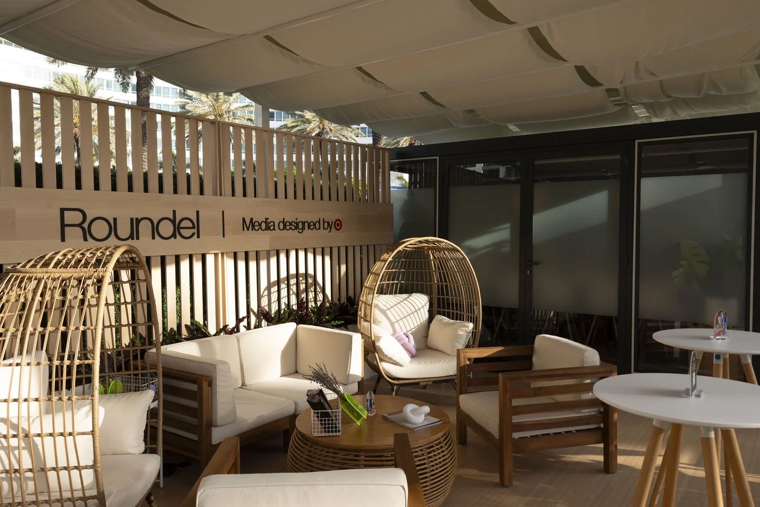 Outdoor seating area with modern wicker furniture, including a curved hanging chair with cushions, a white sofa, and wooden chairs, under a shaded canopy. There are tables with decorative items and a wooden fence with a sign reading 'Roundel | Media designed by' against a backdrop of palm trees and office buildings.