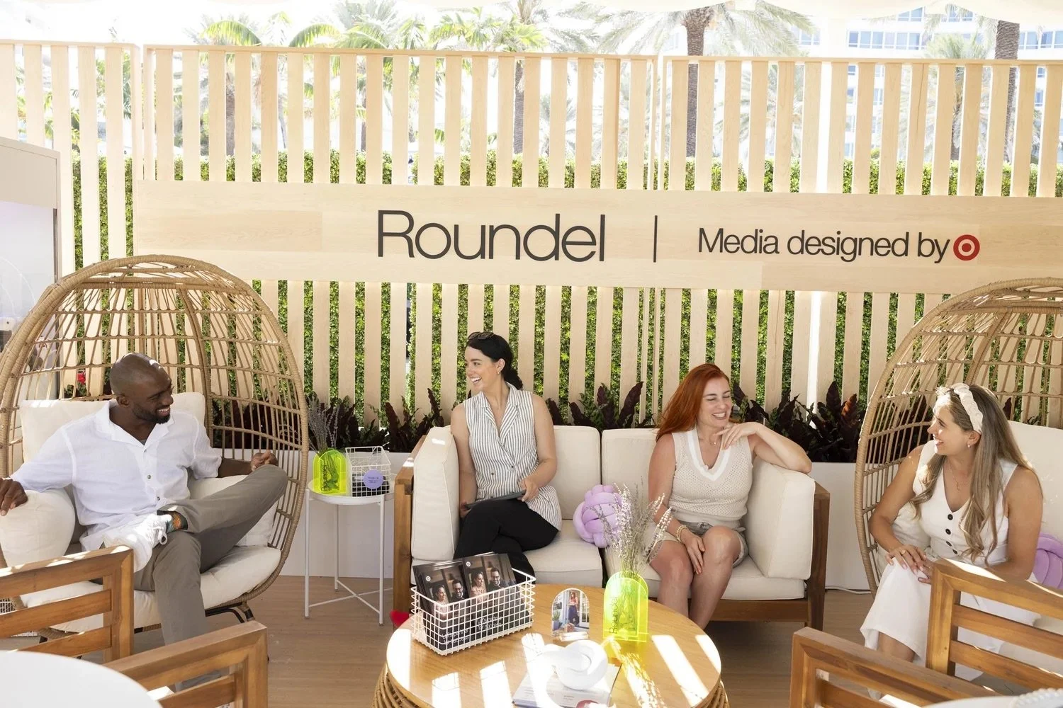 Four people sitting on a white couch and wicker chairs at an outdoor social event, with a wooden background displaying the words 'Roundel' and 'Media designed by Target,' surrounded by plants and trees.