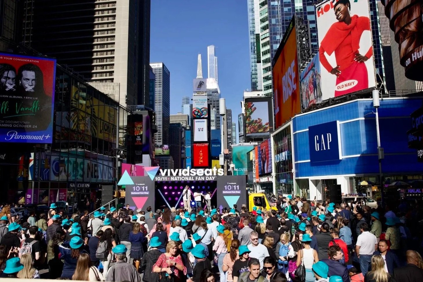 Crowd gathered at an outdoor event in Times Square, with a stage for Vivid Seats' National Fan Day celebration, surrounded by large digital billboards and tall buildings.