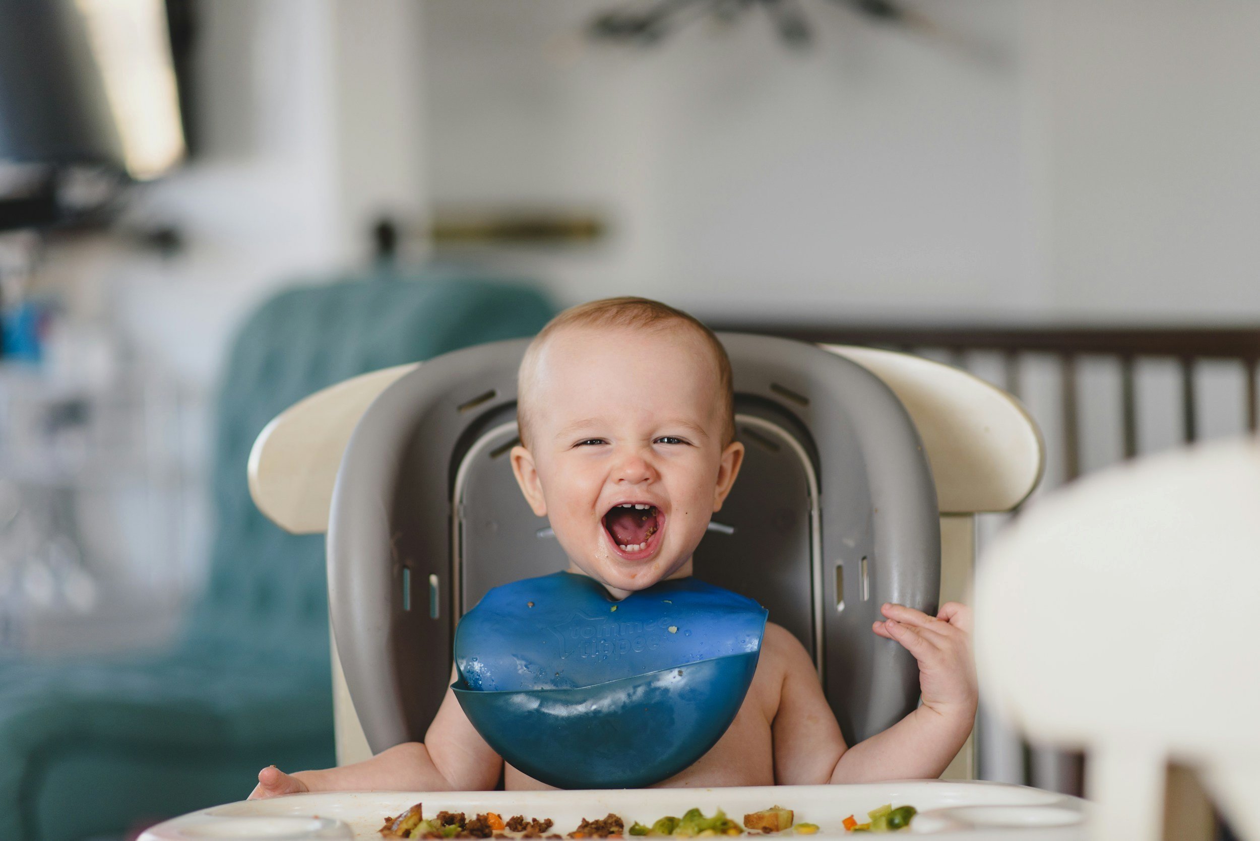 Smiling baby in a high chair with a blue bib, surrounded by food on the tray.