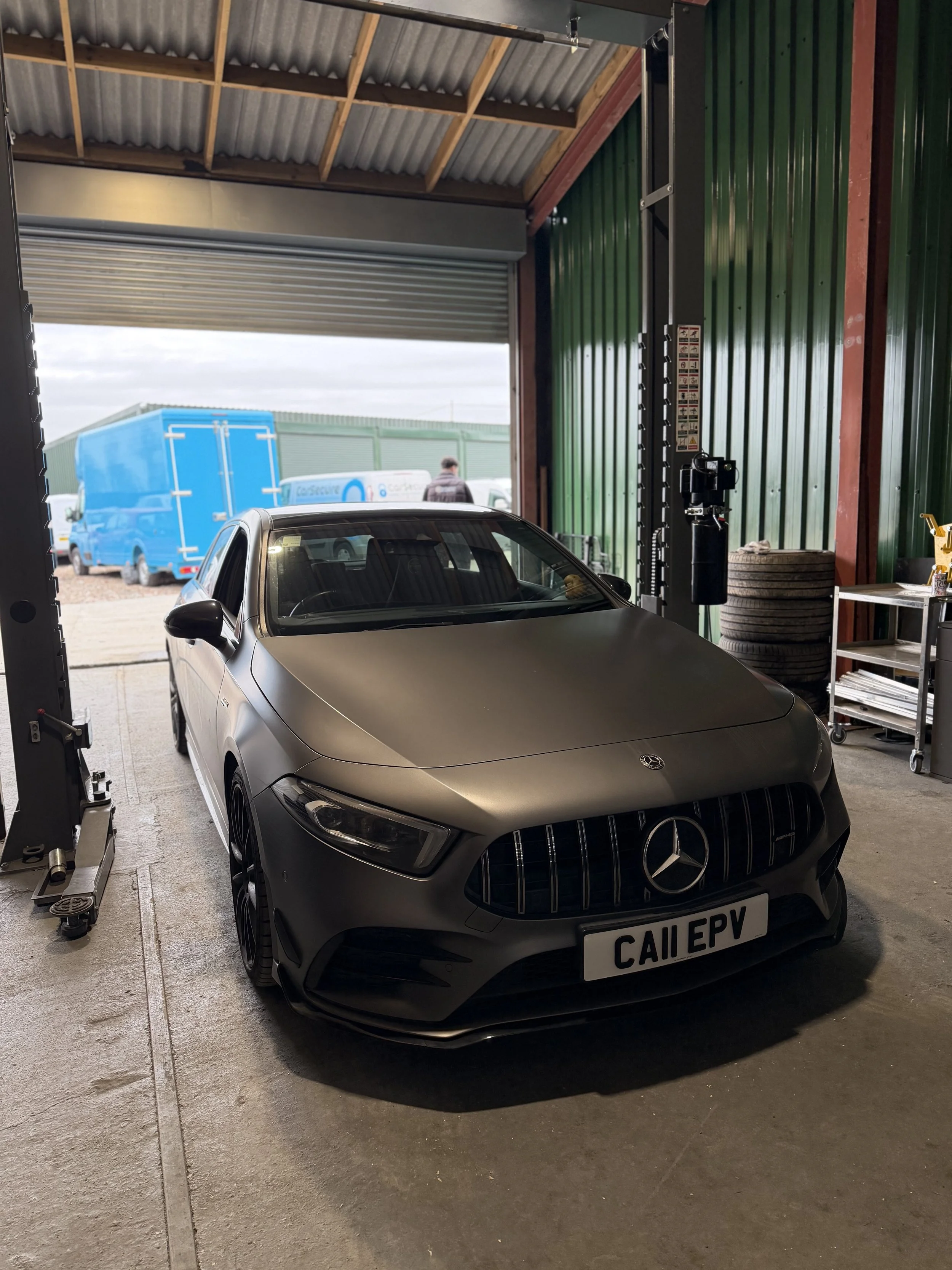 A matte gray Mercedes-Benz car inside a garage with an open roll-up door, showing a blue truck and a person outside.