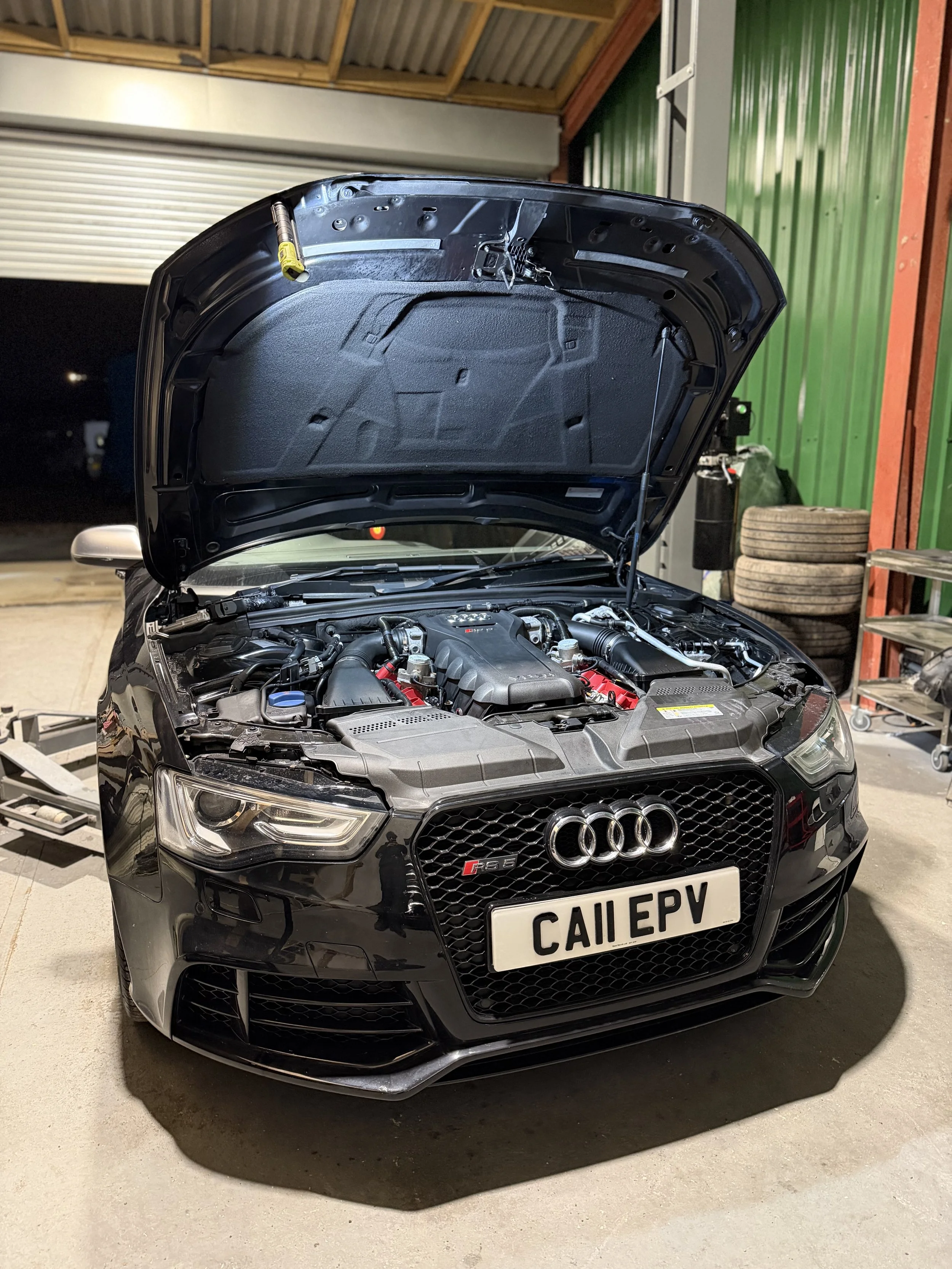 A black Audi car with its hood open inside a garage with green walls, tires stacked in the background, and various tools and equipment nearby.