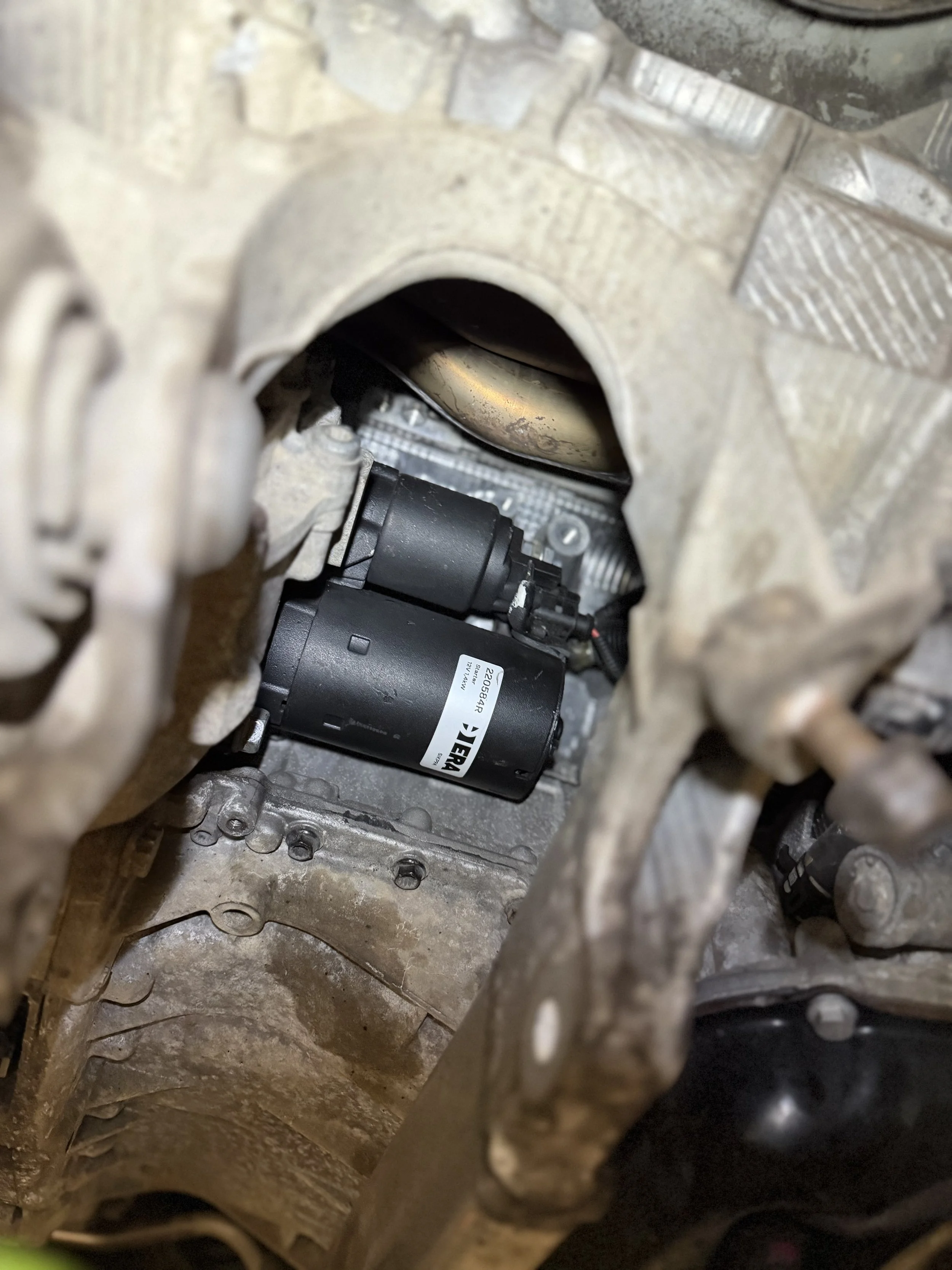 Close-up view of a car engine showing a black electric motor or pump installed inside the engine bay, surrounded by metal components and parts of the engine block.