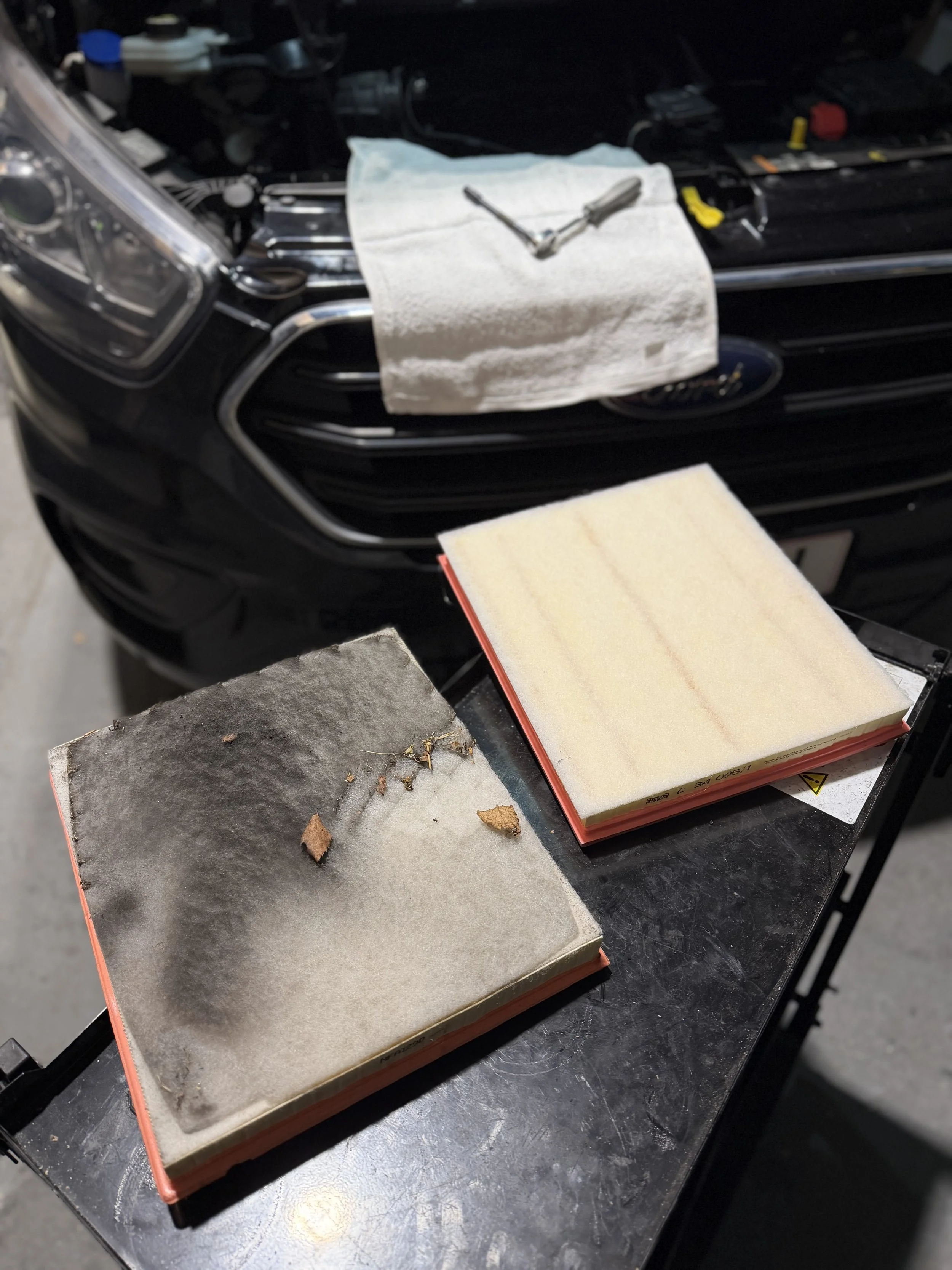 Dirty and clean air filters on a workbench in front of a black vehicle engine, with a towel, screwdriver, and other tools nearby.