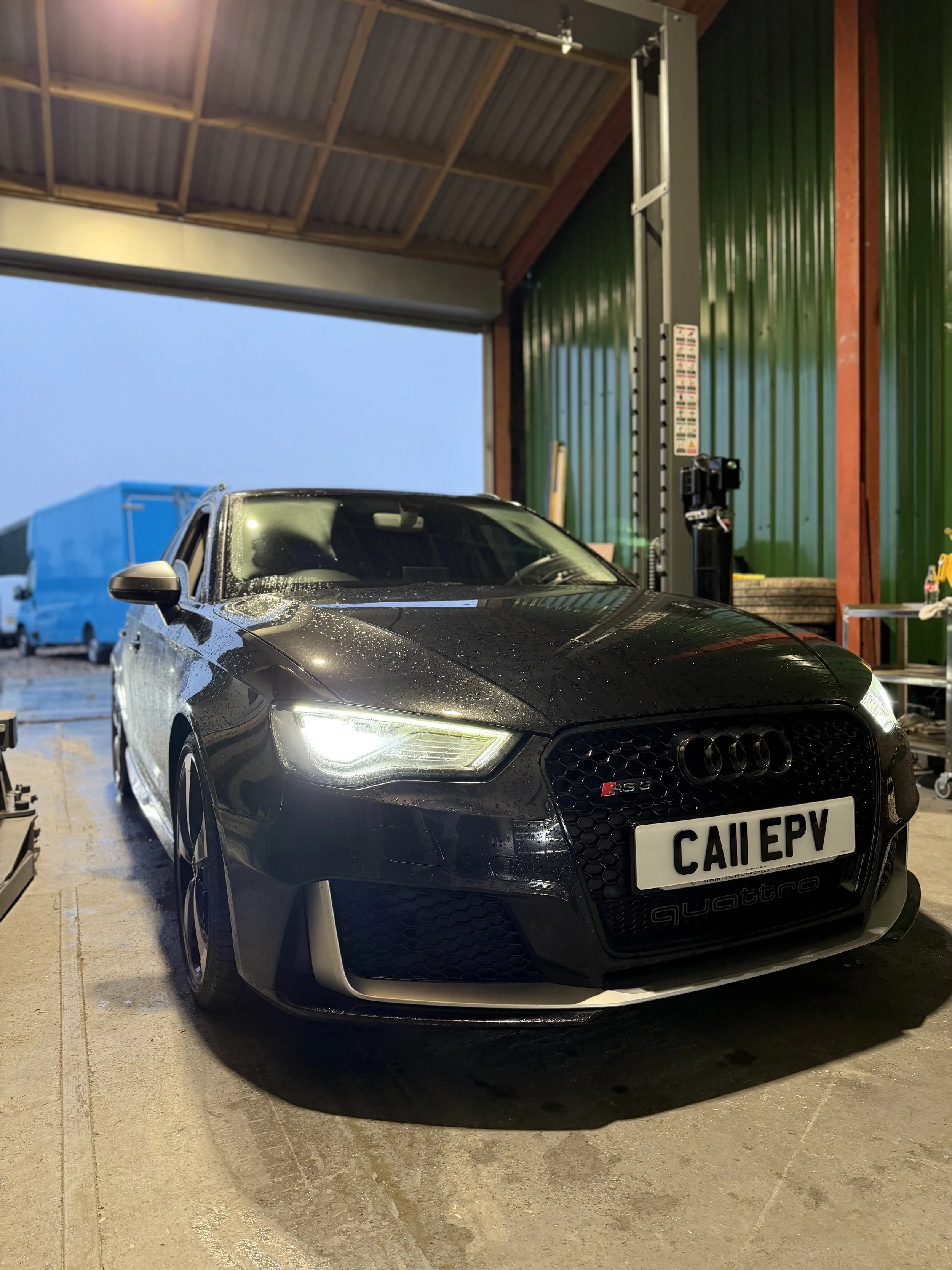 A black Audi RS3 parked inside a garage, wet from rain, with a green wall and tools on a cart in the background.