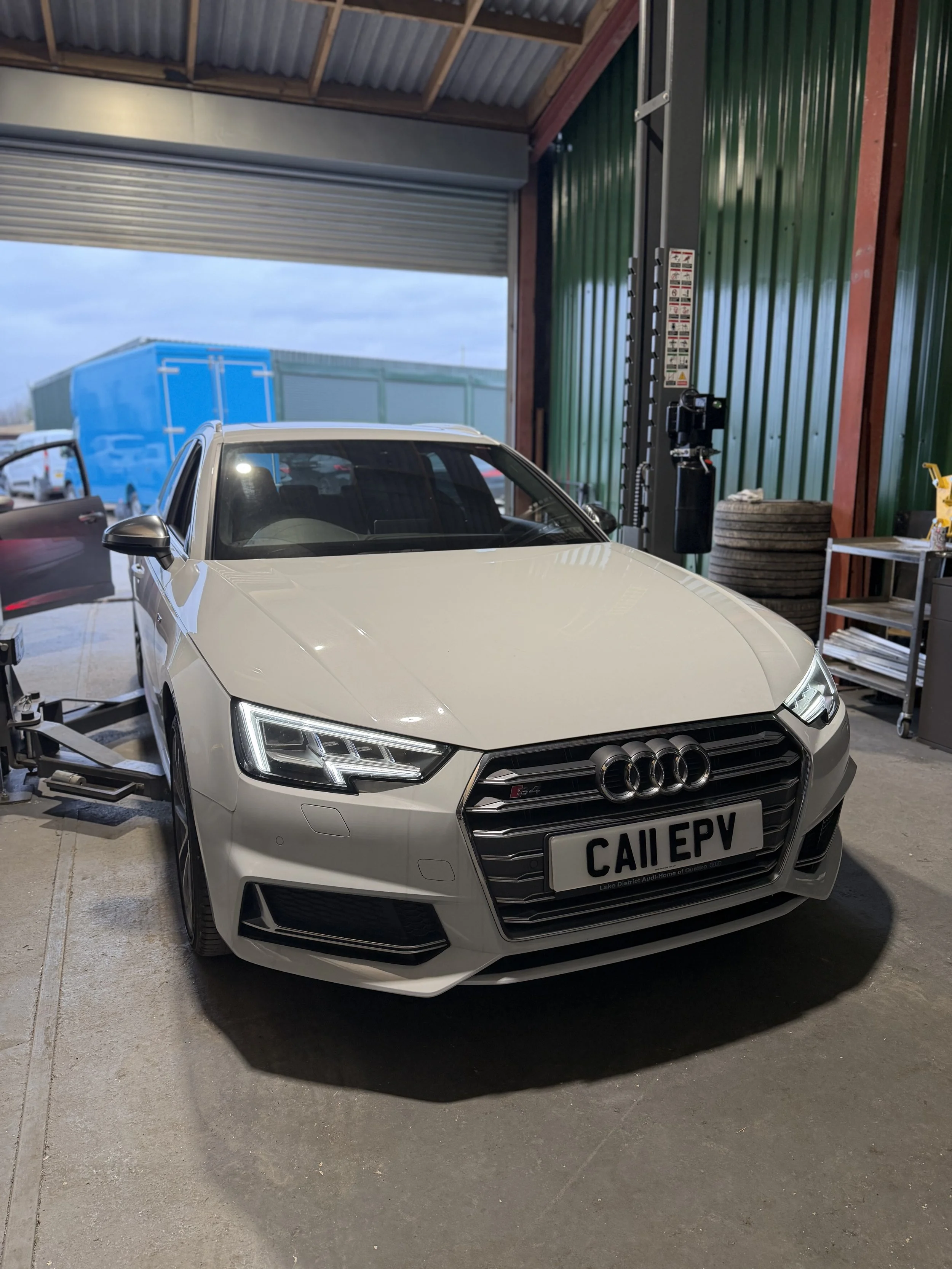 A white Audi S4 sports car with a custom front license plate reading 'CAII EPV', parked inside a garage next to a machine, with an open garage door revealing a blue truck outside.