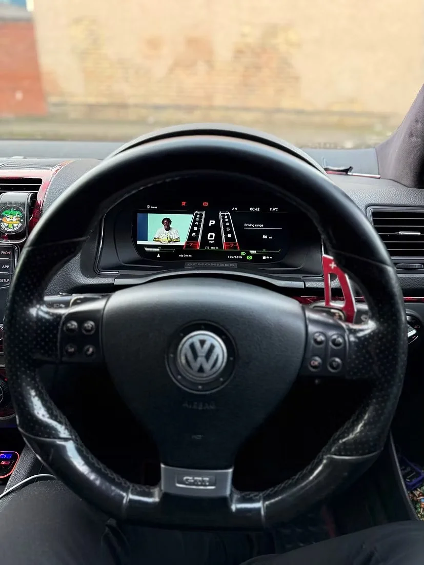 The image shows the interior of a Volkswagen car from the driver's seat perspective. The dashboard displays digital gauges and a small screen, and there's a TV show or movie playing on the digital display. The steering wheel has the Volkswagen logo.