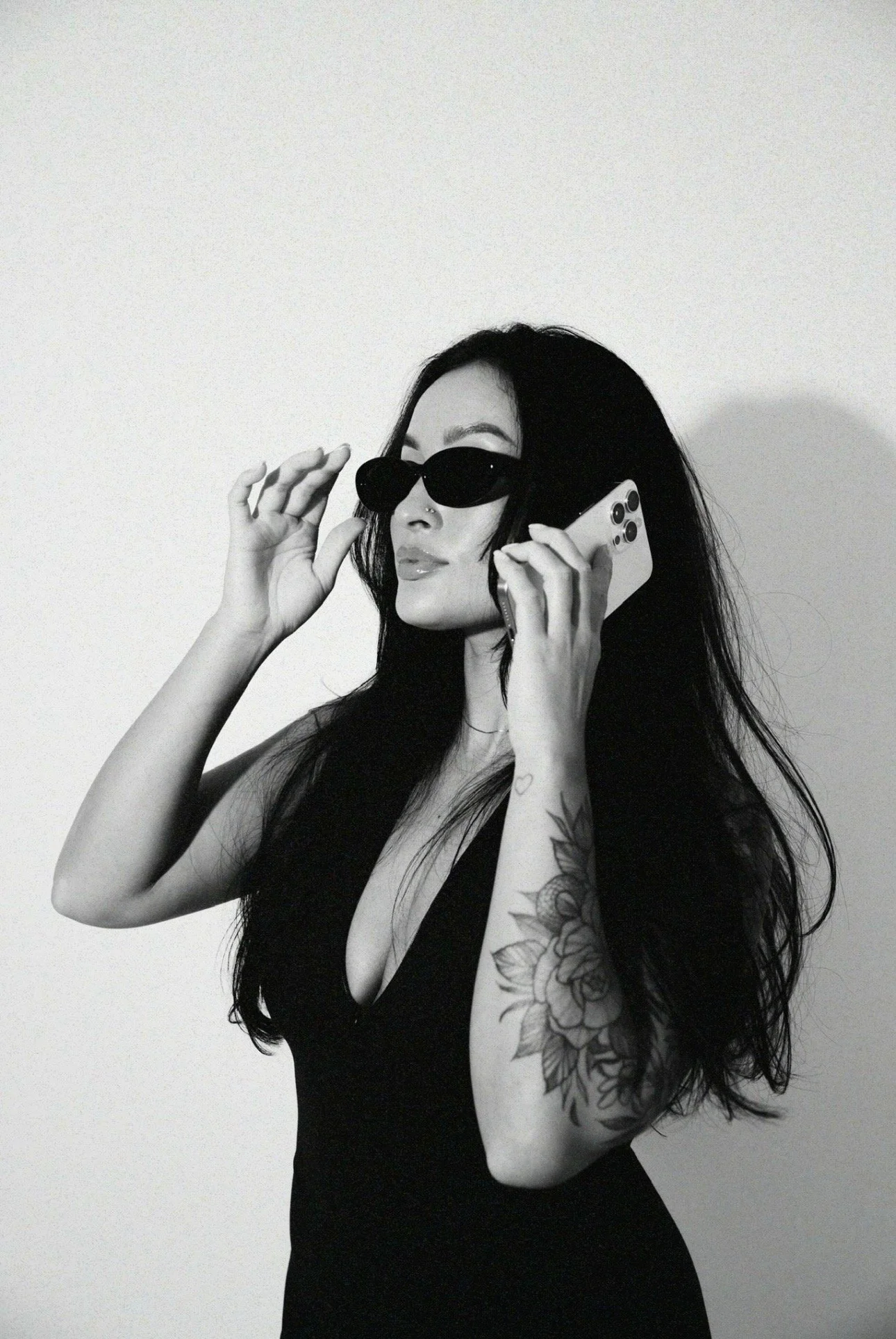 A woman with long dark hair, wearing sunglasses and a sleeveless black top, holding a phone to her ear, standing against a plain background.
