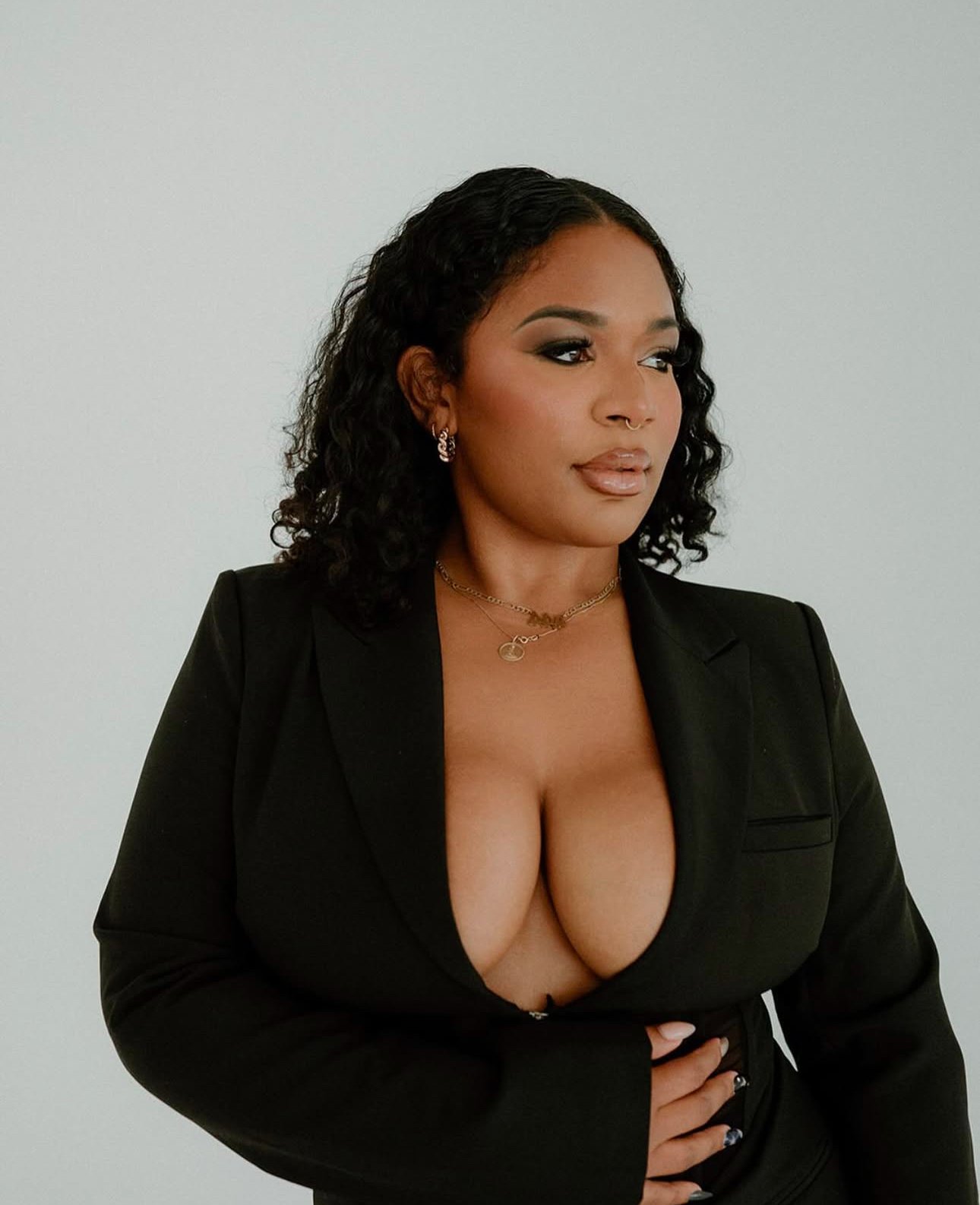 A woman with curly black hair, wearing a black blazer and revealing top, standing against a plain white background.