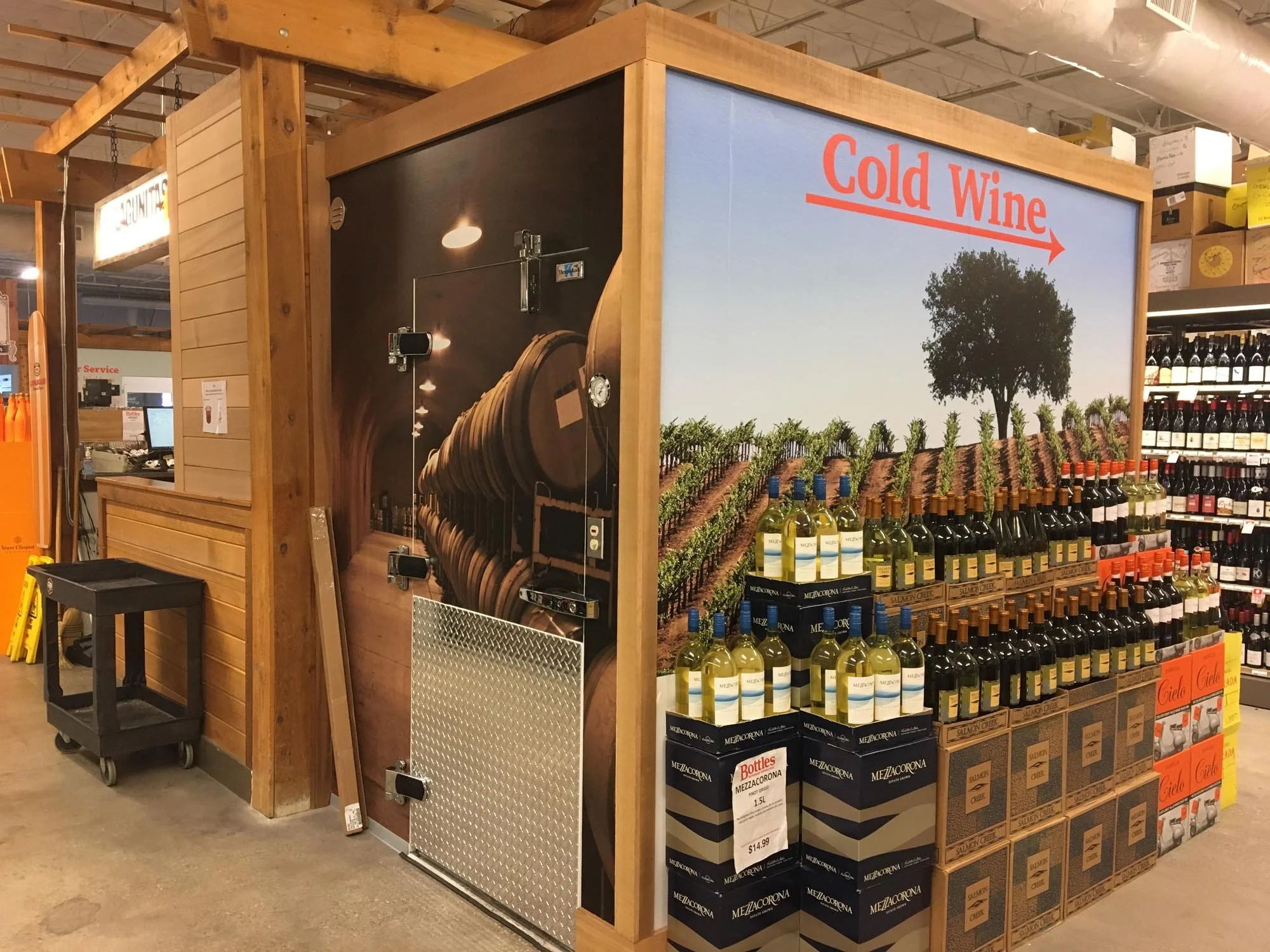 Wine display inside a store with a large sign reading 'Cold Wine' pointing to the right, and bottles of white and red wine lined up on a shelf in front of a vineyard backdrop.