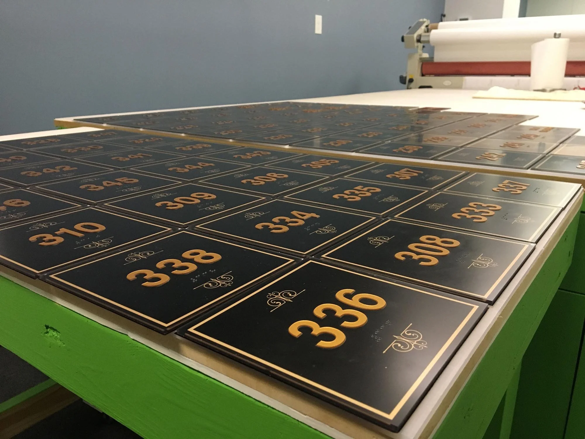 Black plaques with gold numbers and decorative details arranged on a green table in a workshop.