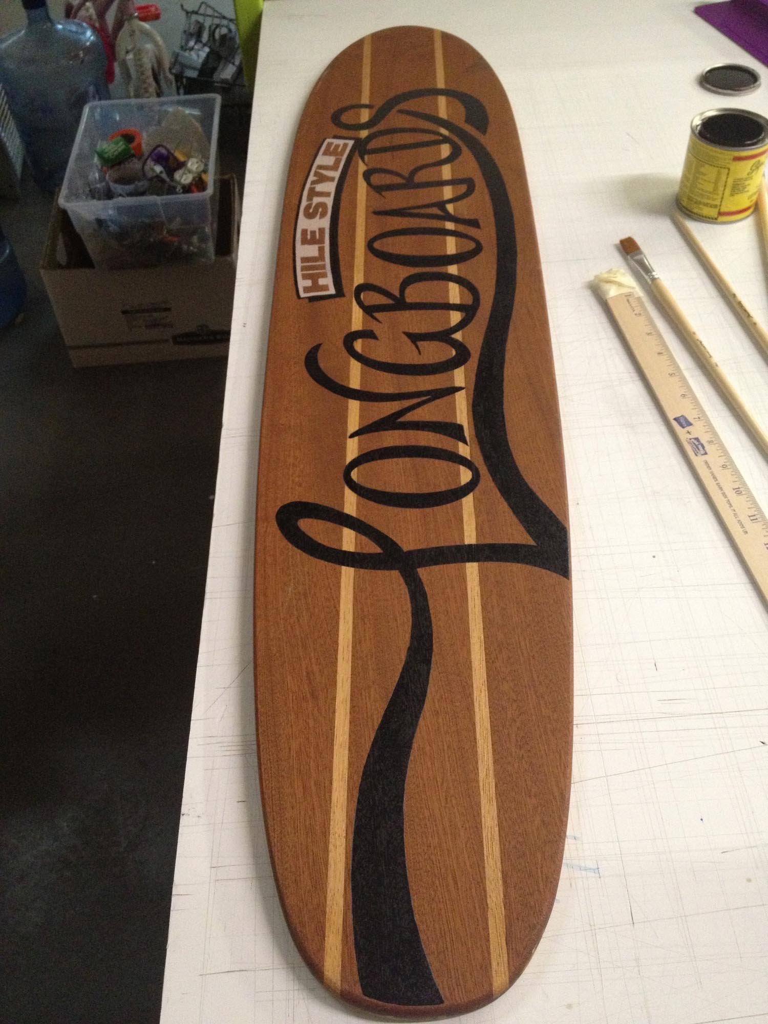A wooden skateboard deck with the words 'Longboards' and 'Hile Style' painted on its surface, laid on a white work surface with paint brushes and a can of paint nearby.