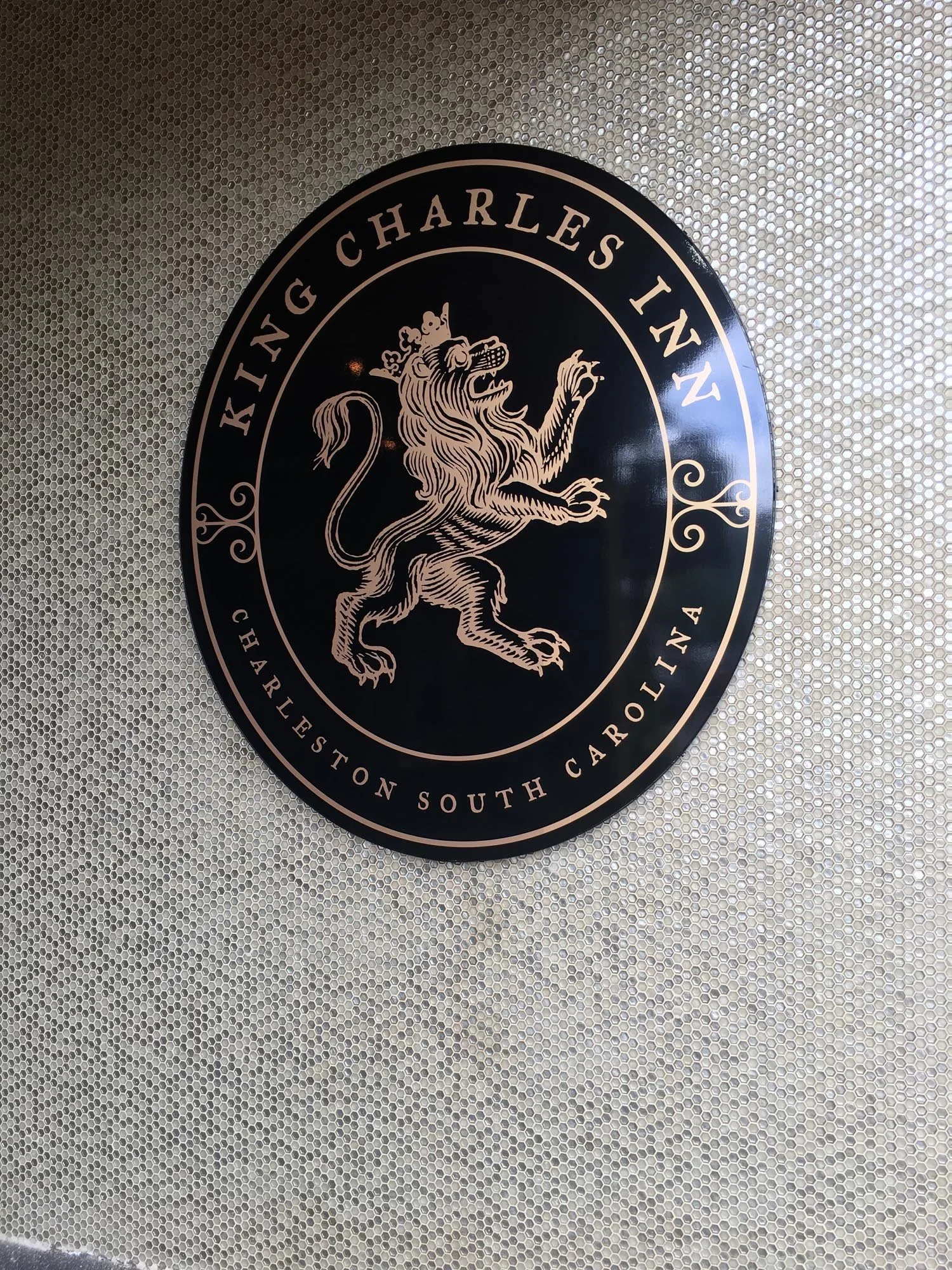 Round black logo with a lion wearing a crown, surrounded by text that reads 'King Charles Inn' and 'Charleston South Carolina' on a metallic, perforated background.