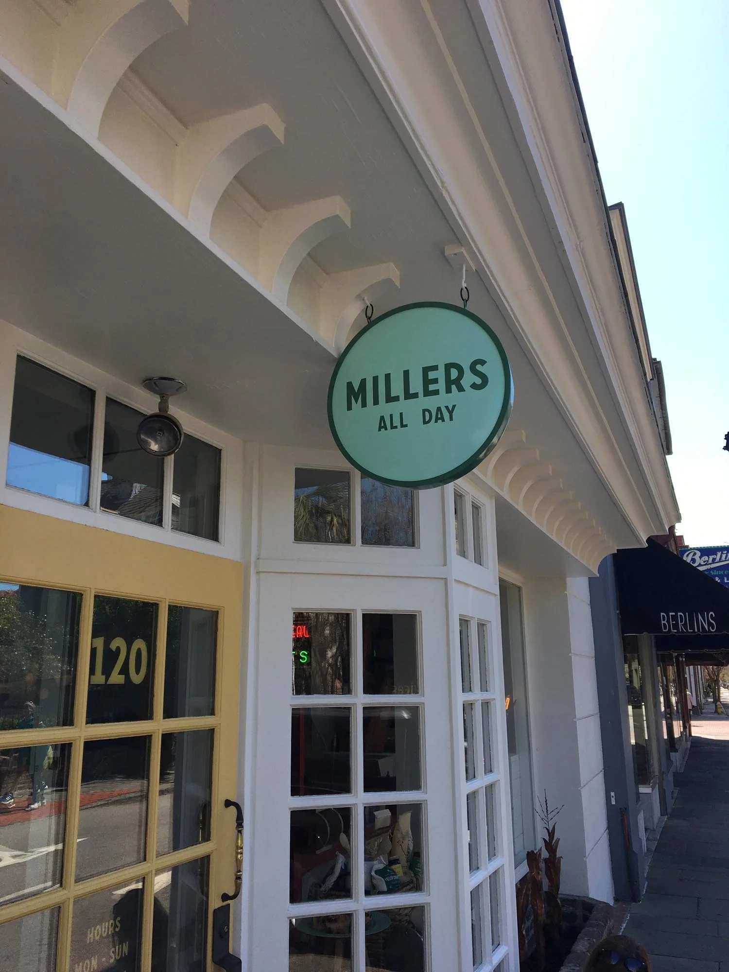 A circular sign hanging above a storefront reads 'Millers All Day.' The storefront has a yellow door and white-framed windows, with a black sign to the right reading 'Berlins.'