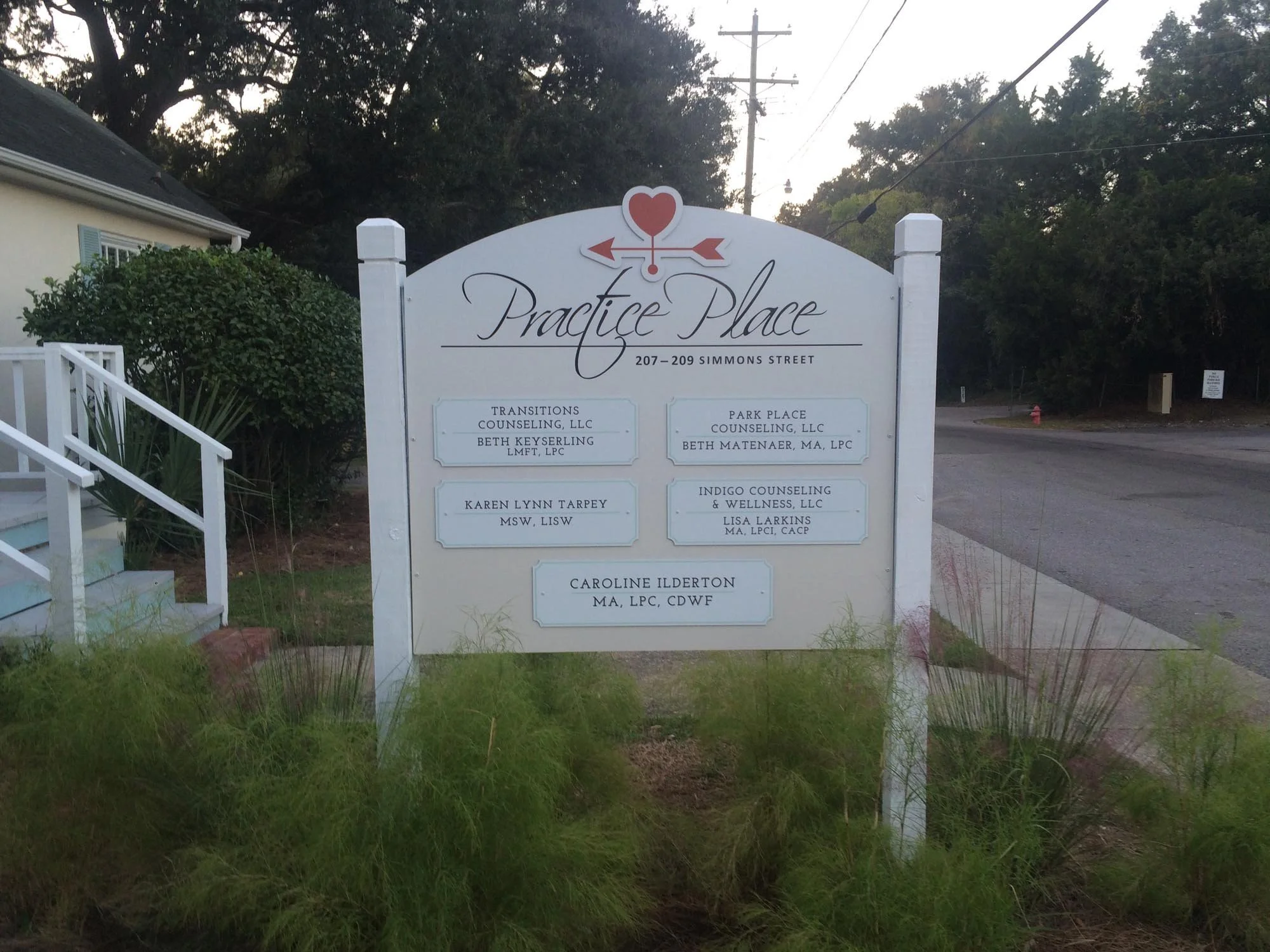A white sign with black and decorative text reading 'Practice Place, 207-209 Simmons Street'. The sign lists several names and credentials of professionals including counselors and therapists, and is situated outdoors near a sidewalk, house, bushes, 