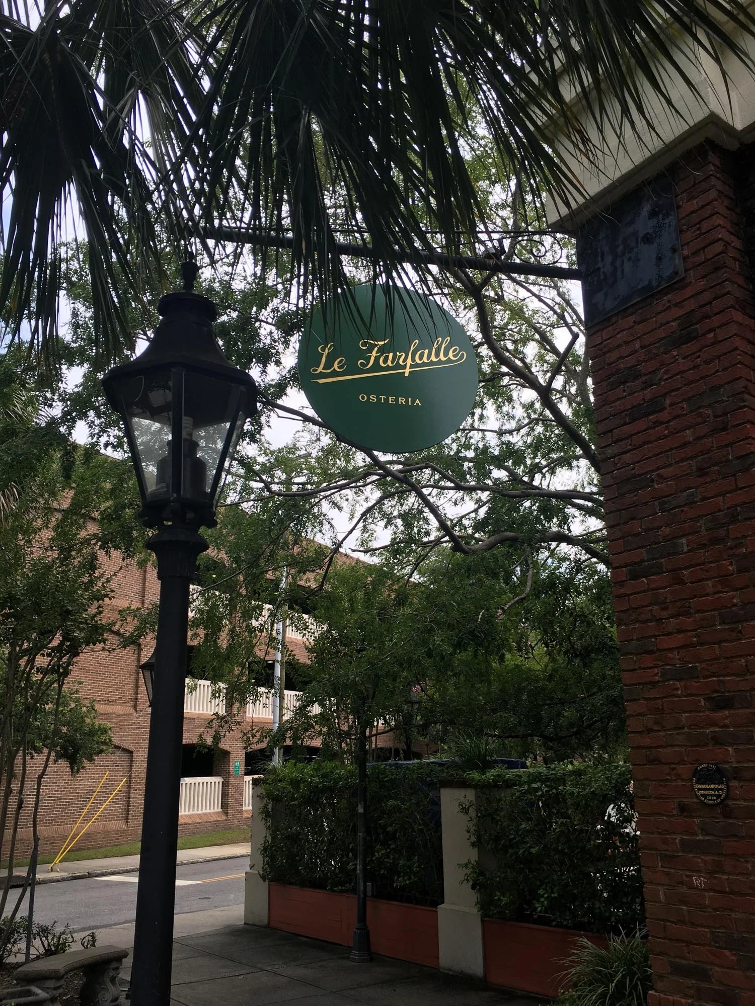 Street view showing the sign of Le Farfalle Osteria hanging above sidewalk, with trees, brick building, and street in the background.