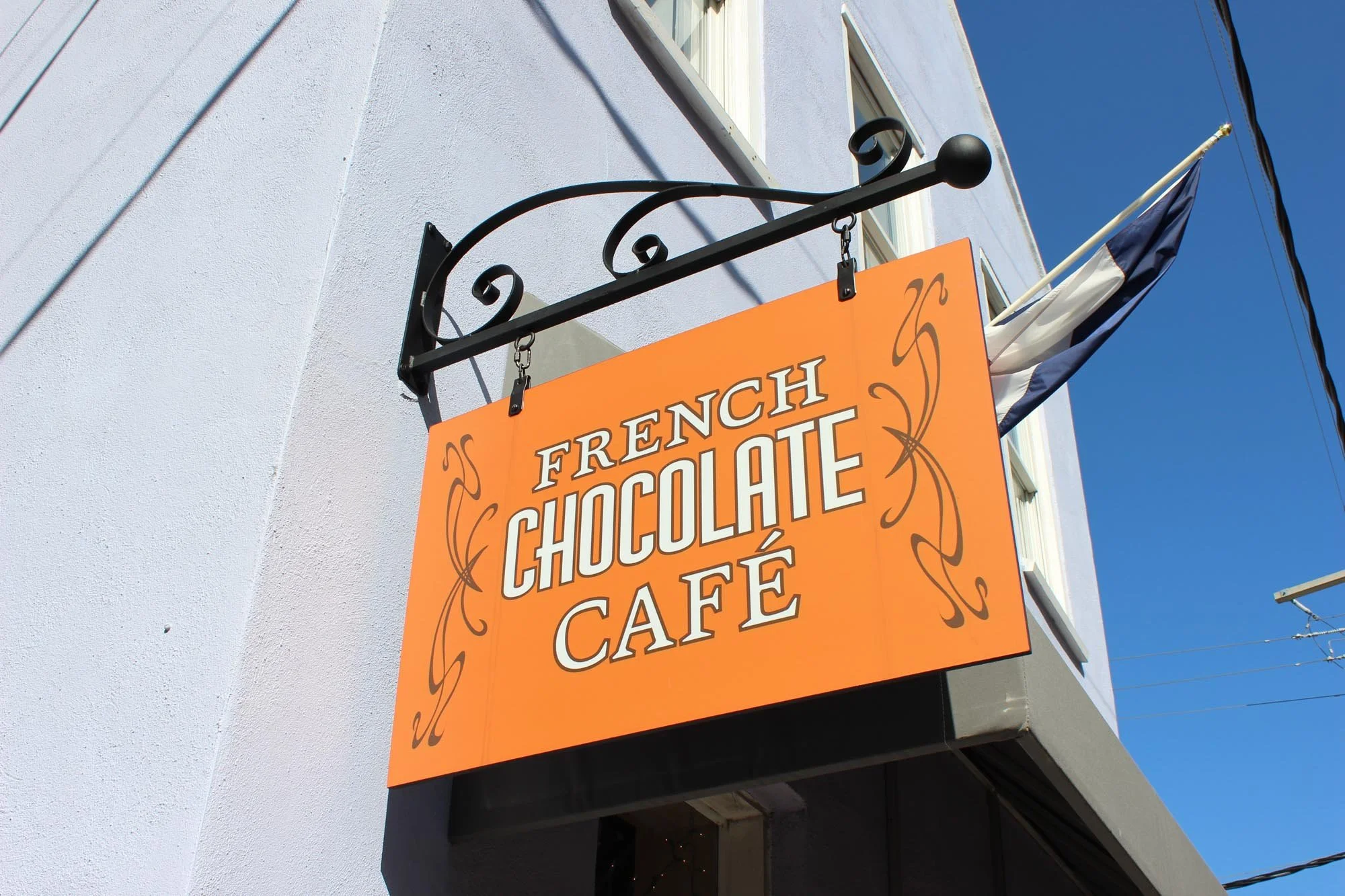 Orange sign that reads 'French Chocolate Café' hanging outside a building with a flag and a clear blue sky.