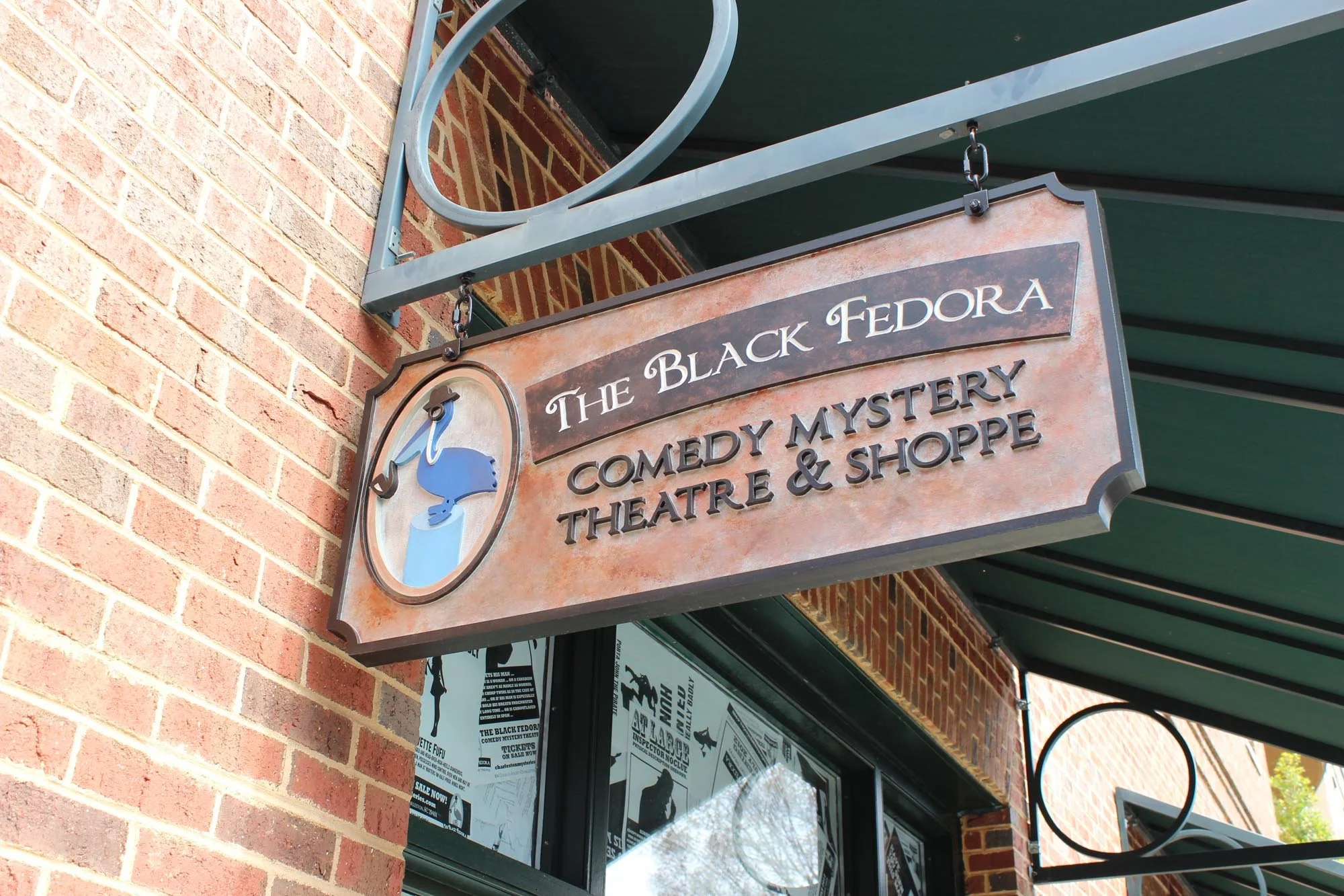Outdoor sign for The Black Fedora Comedy Mystery Theatre & Shoppe hanging from a metal arm against a brick wall with a green awning above.