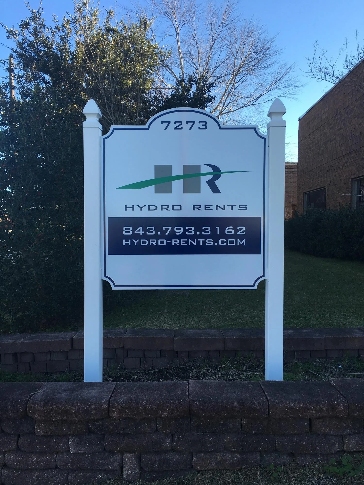 A white signboard with the logo and contact information for Hydro Rents, located on a sidewalk near some bushes and trees with a brick building in the background.