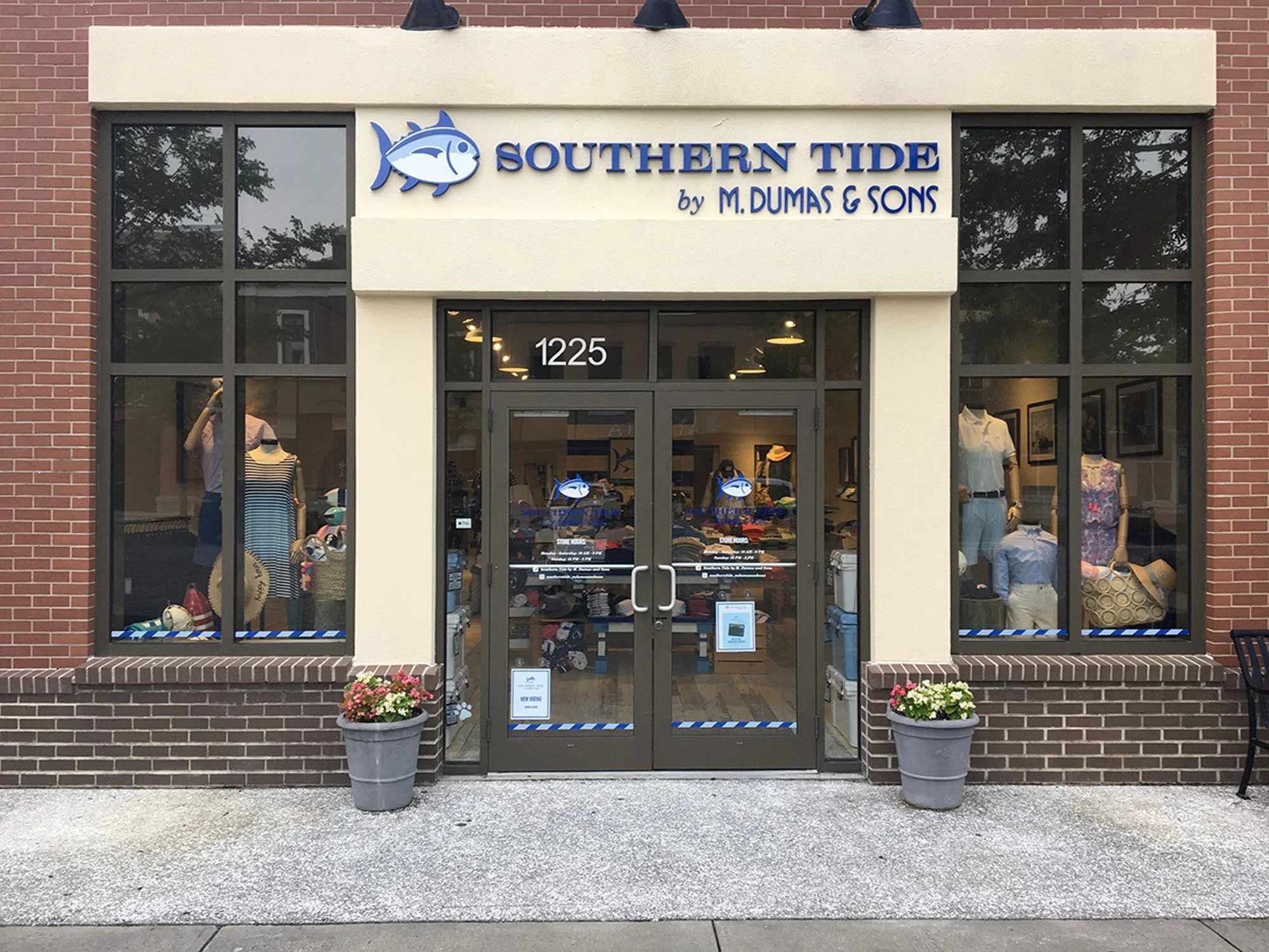 Storefront of Southern Tide clothing store with large glass windows displaying mannequins dressed in casual apparel, the store's name in blue, with a fish logo, and two potted flowers outside.