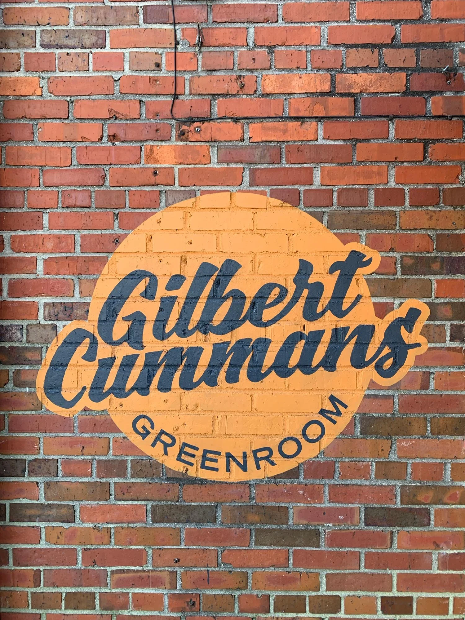Sign painted on a brick wall that says 'Gilbert Cummings Greenroom' with the words in black inside an orange circle.