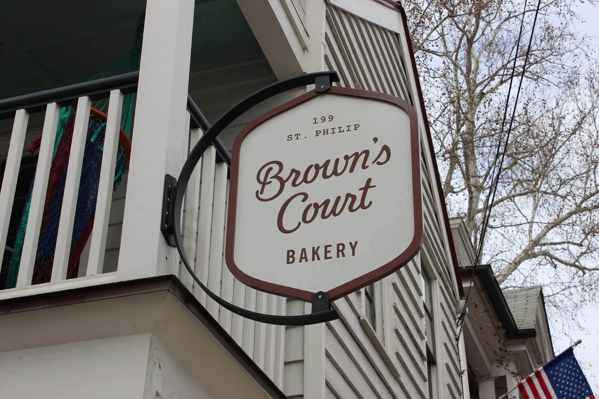 A sign for Brown's Court Bakery hanging on a building's exterior with trees and a flag in the background.