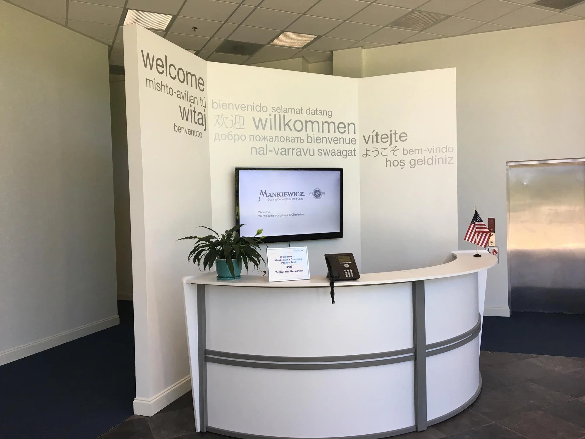 A modern reception desk with a potted plant, a telephone, a computer monitor, and small American flags. Behind the desk is a wall with multilingual welcome messages and a mounted screen displaying a title that appears to relate to coaching concepts. 