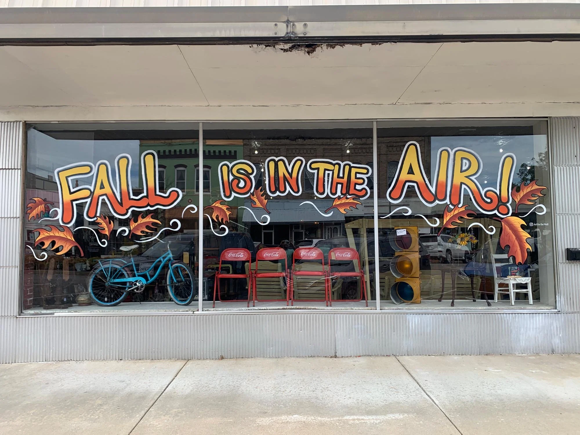 Storefront window with fall-themed decorations and a sign that says 'Fall is in the Air!' in colorful letters, surrounded by painted autumn leaves, a blue bicycle, red Coca-Cola chairs, a yellow traffic light, and a white chair inside.
