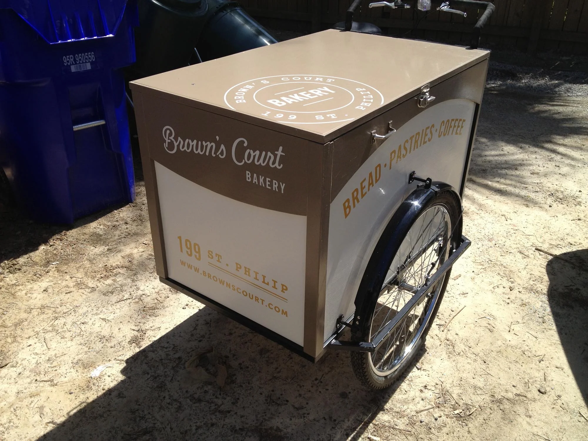 A bakery cart on wheels with signs for Brown's Court Bakery, featuring bread, pastries, and coffee, positioned outdoors on a dirt ground.