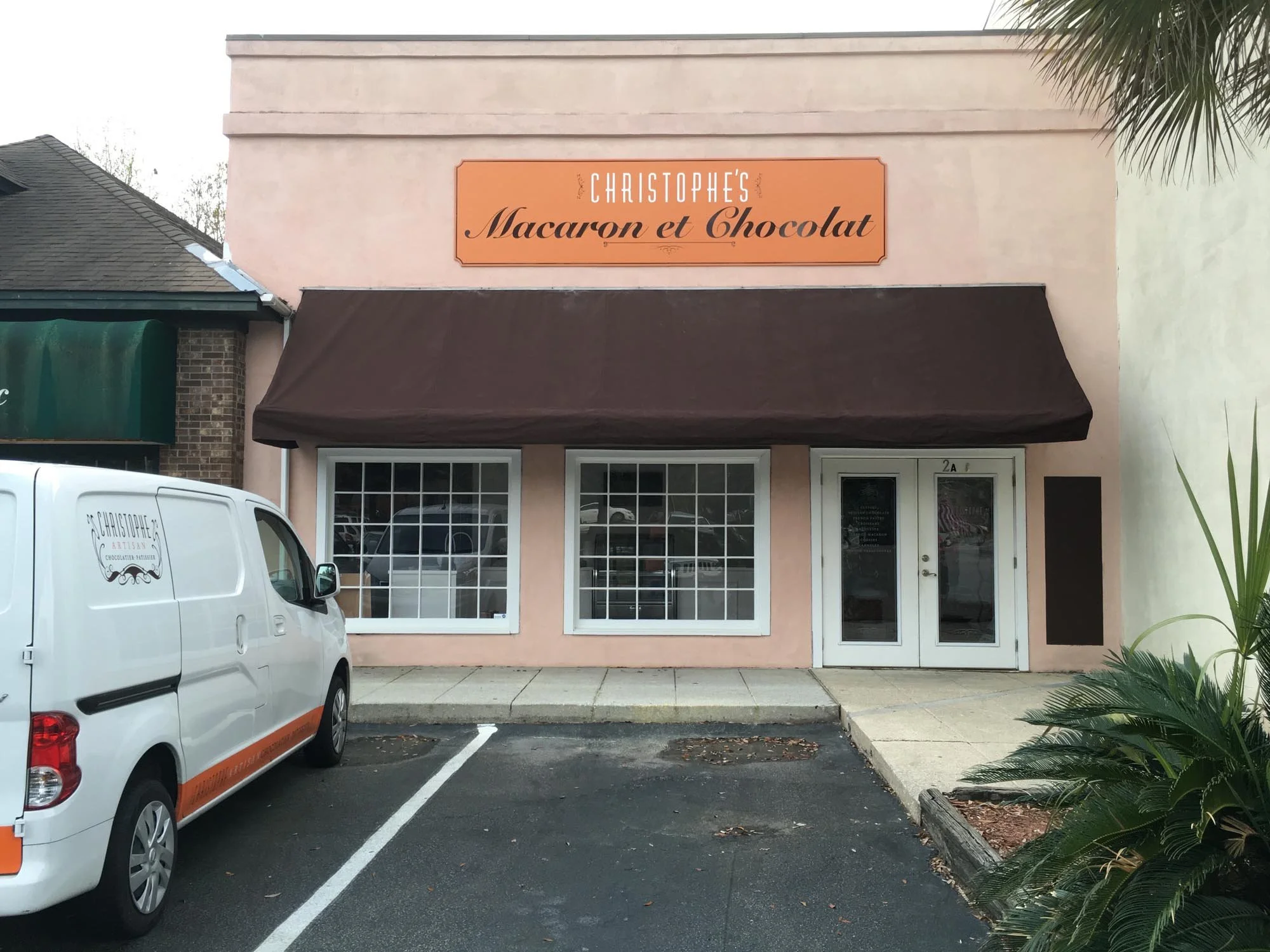Front of a bakery with a sign reading 'Christophe's Macaron et Chocolat,' two large windows, a glass door, a white van with the bakery's logo, and parking spaces in front.