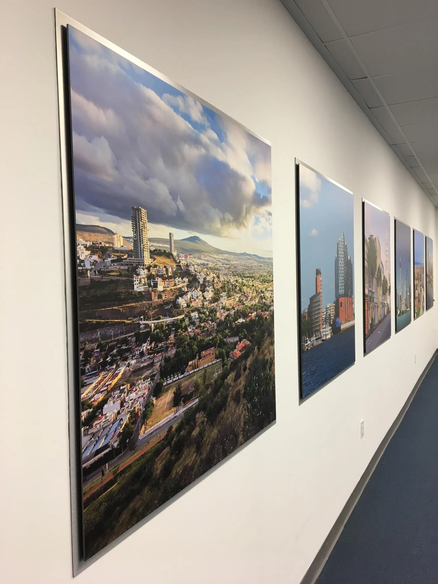 A series of framed cityscape photographs displayed on a white gallery wall inside a building, depicting urban skyline scenes with buildings, hills, and sky in the background.