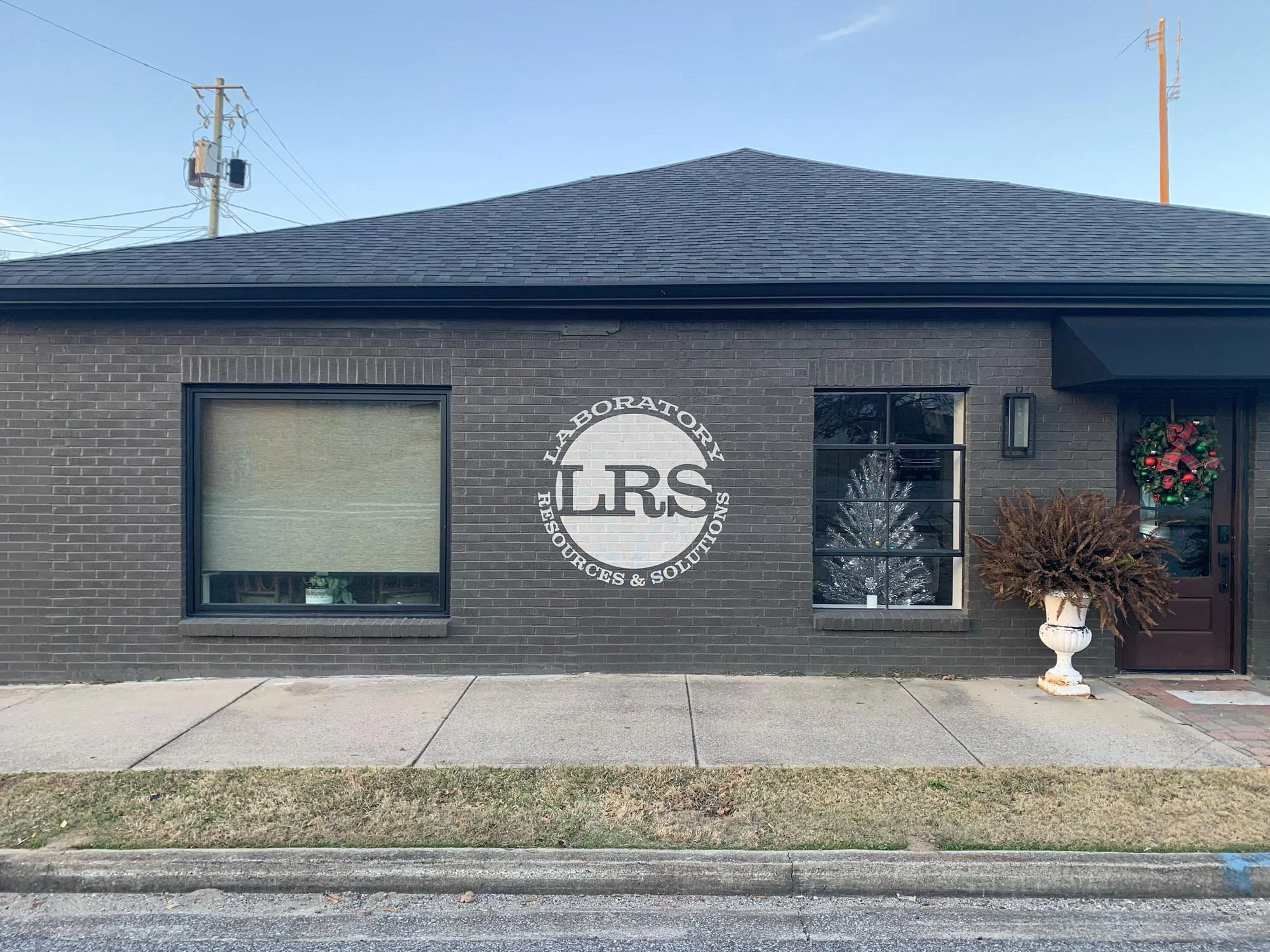 Black brick building with sign reading "LRS Laboratory Resources & Solutions" painted on exterior wall, large window with green plant inside, Christmas wreath on door, potted plant outside, sidewalk in front, and utility poles in background.