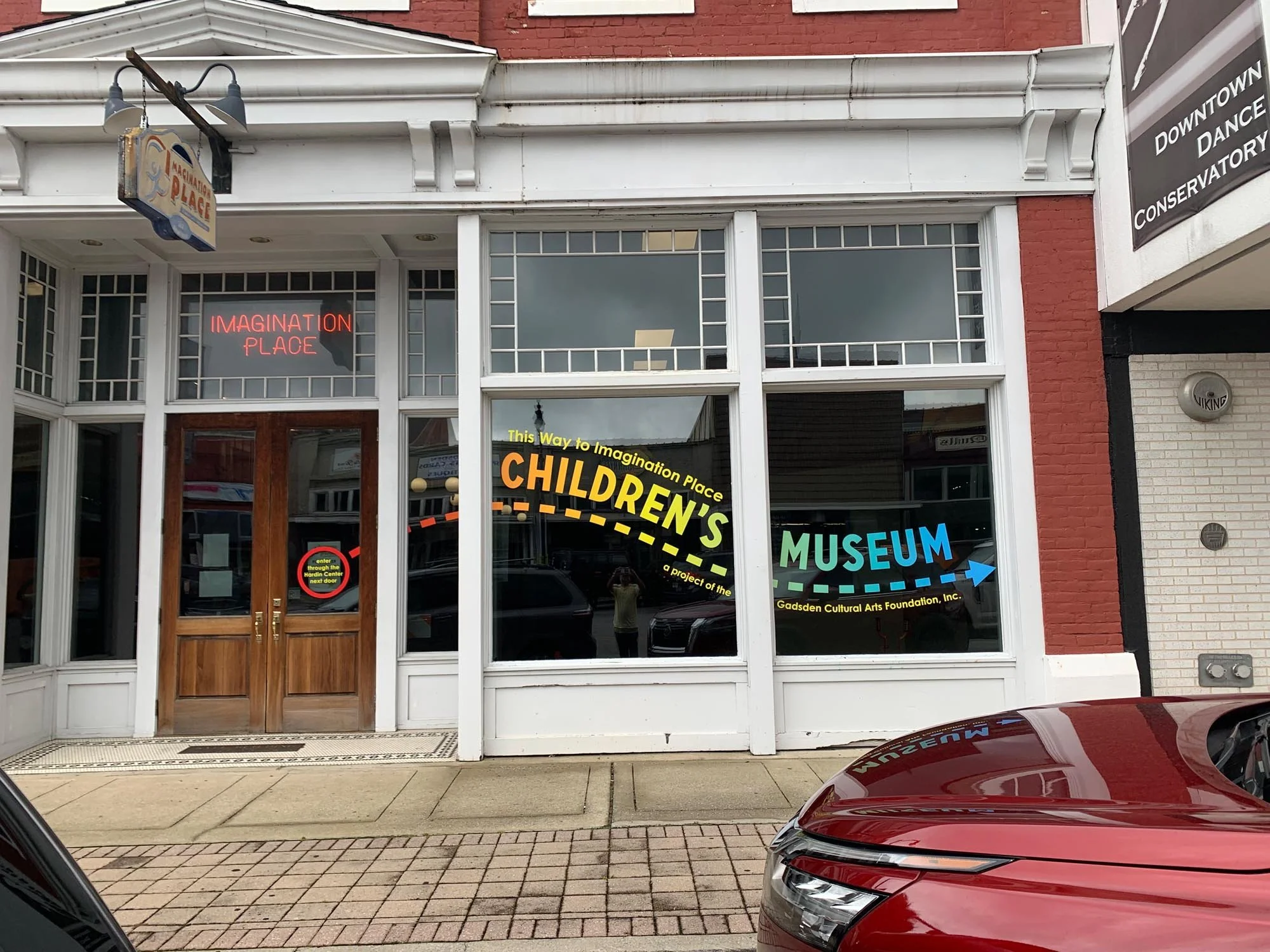 Front view of Imagination Place Children's Museum with large windows, wooden double doors, and colorful signs indicating the museum's name and features. A red car is parked in front on the street.