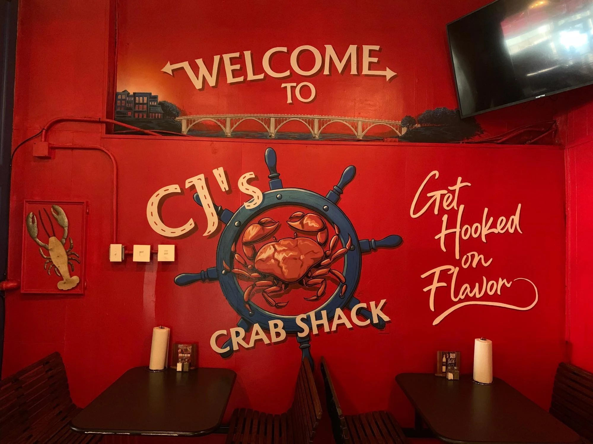 Red wall inside a seafood restaurant with a mural featuring a crab at the center of a ship's steering wheel. Above, a sign reads 'Welcome to CJ's Crab Shack.' To the right, a message says 'Get Hooked on Flavor.' There are tables with condiments, a pa