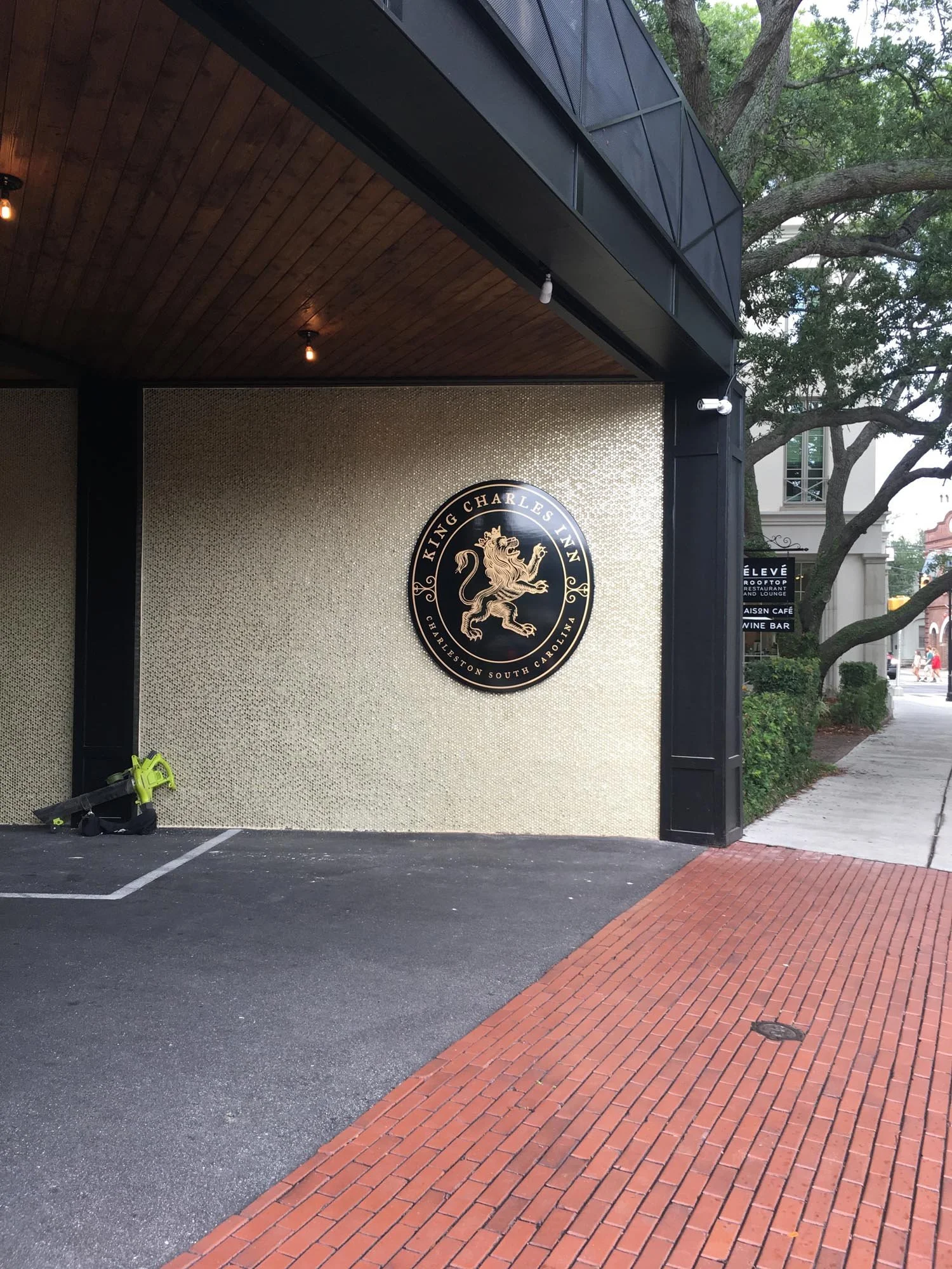 Exterior of King Charles Inn hotel with beige textured wall, black logo with a lion, and a black metal and wooden ceiling, with trees and a sidewalk in the background.