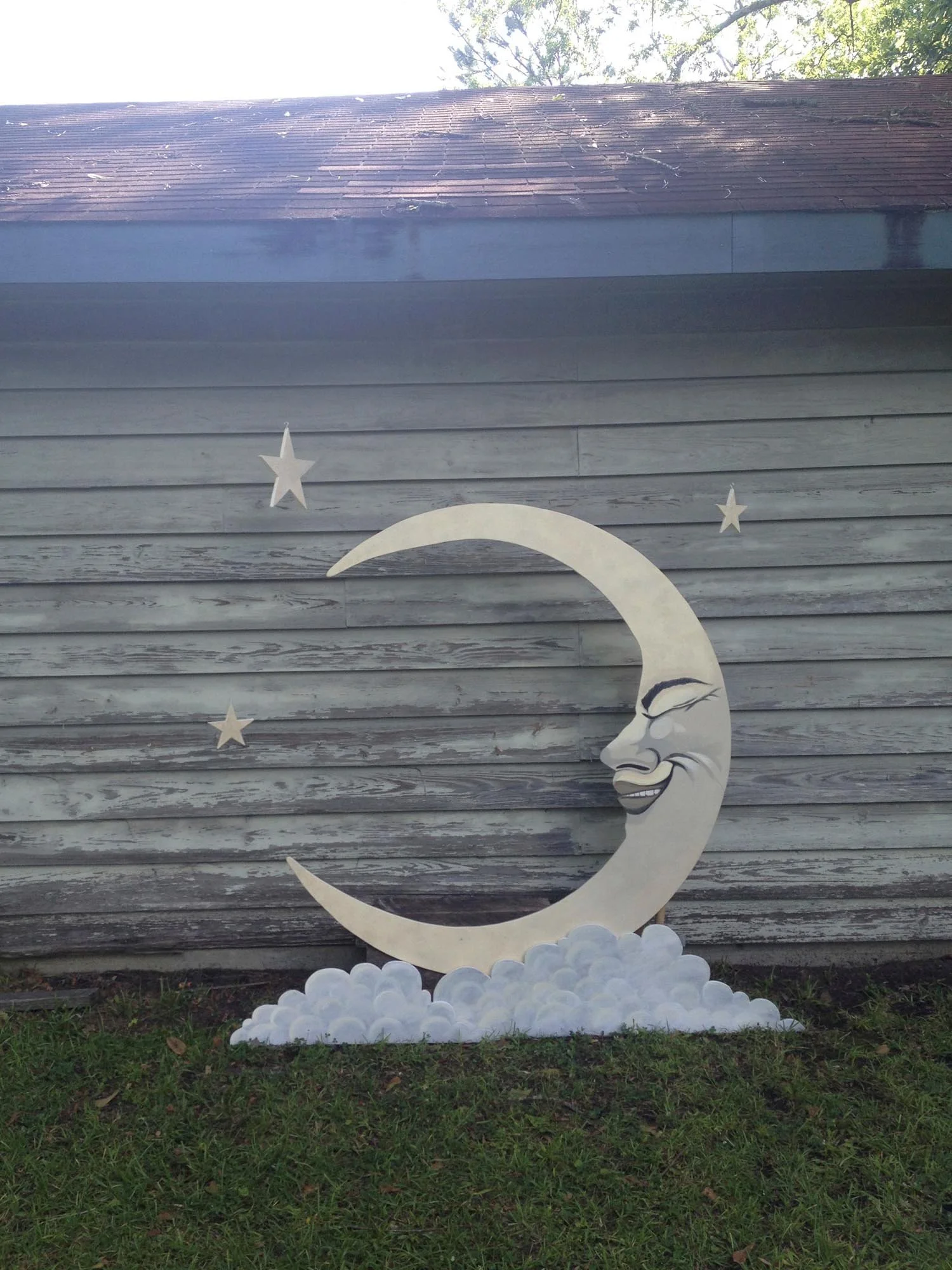 Decorative moon with a face, surrounded by clouds and three stars, mounted on a weathered wooden wall, outdoors.