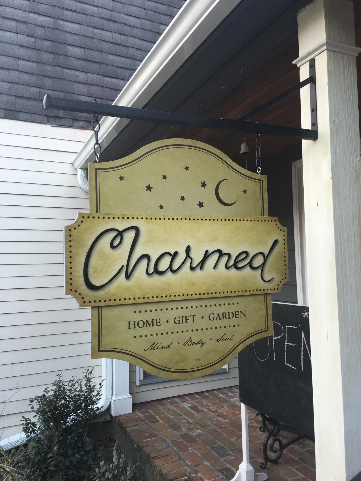 A yellow and black outdoor sign for a store called 'Charmed,' offering home, gift, and garden items, with a moon and stars design and the phrase 'Mind, Body, Soul' at the bottom.