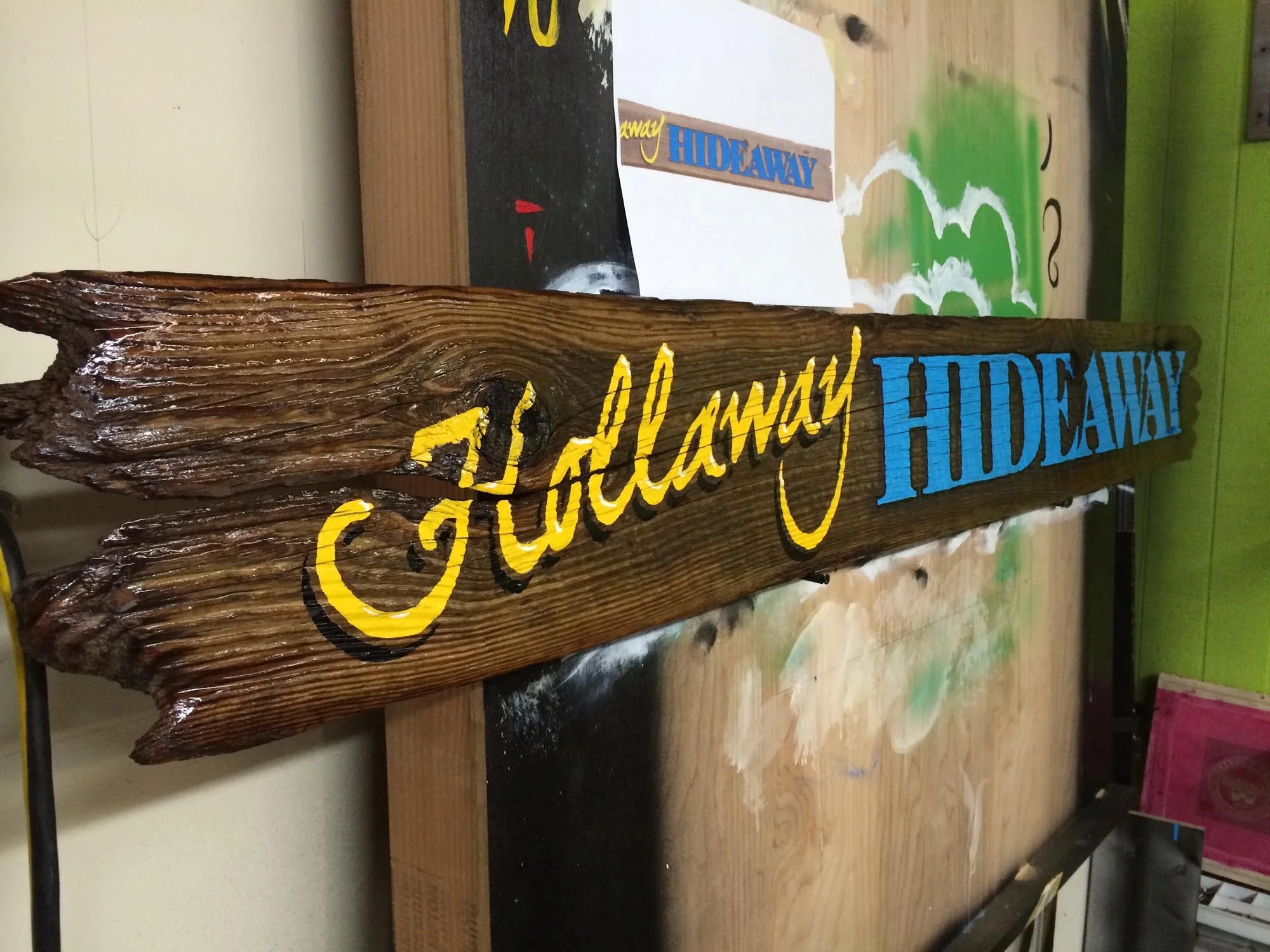 Wooden sign with the words 'Always HIDEAWAY' painted in yellow and blue, resting on a worktable with paint spots.