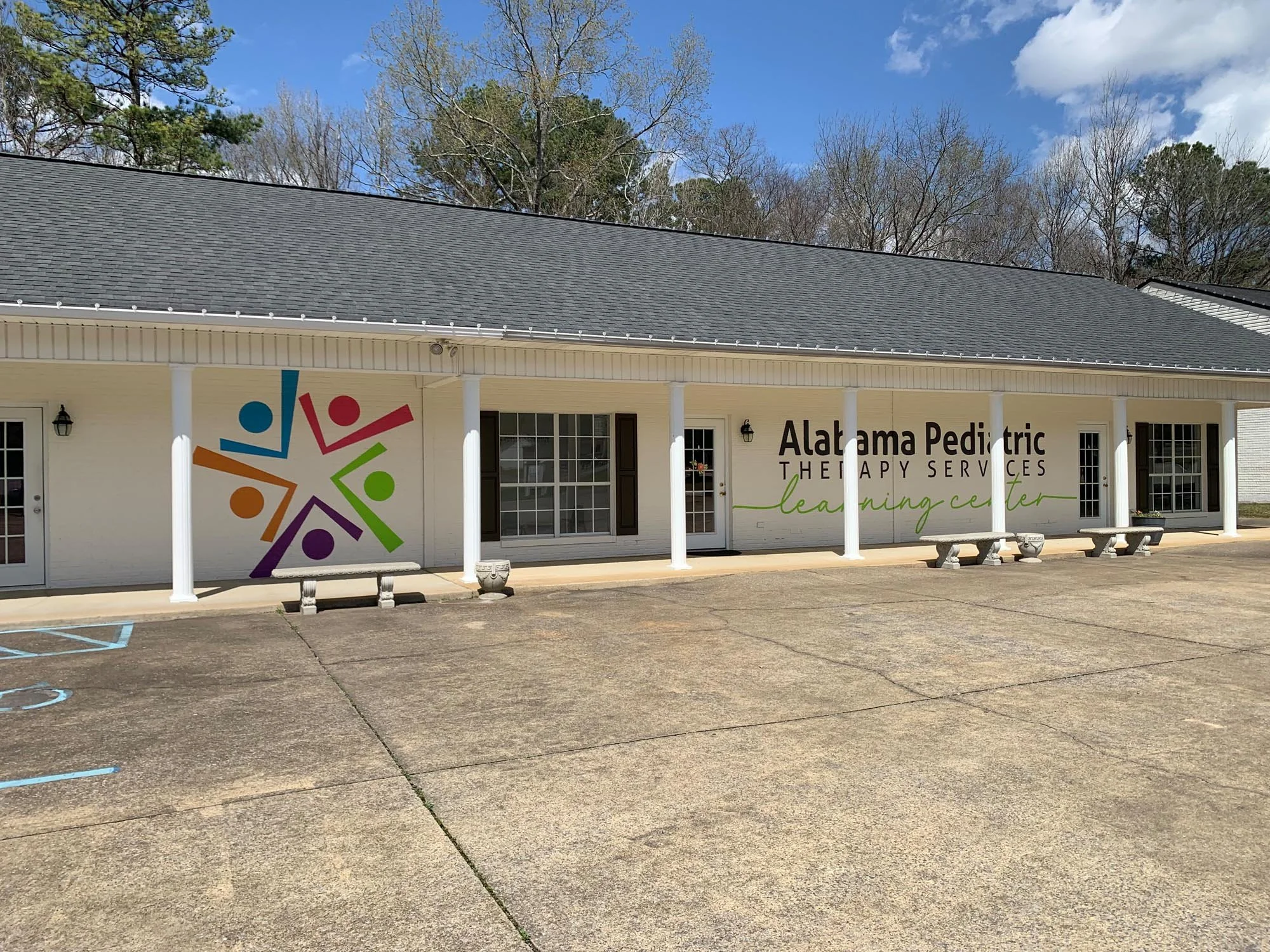 Front view of Alabama Pediatric Therapy Services Learning Center building with a paved parking lot, benches, white exterior walls, black shutters, and a colorful logo featuring abstract human figures holding hands.