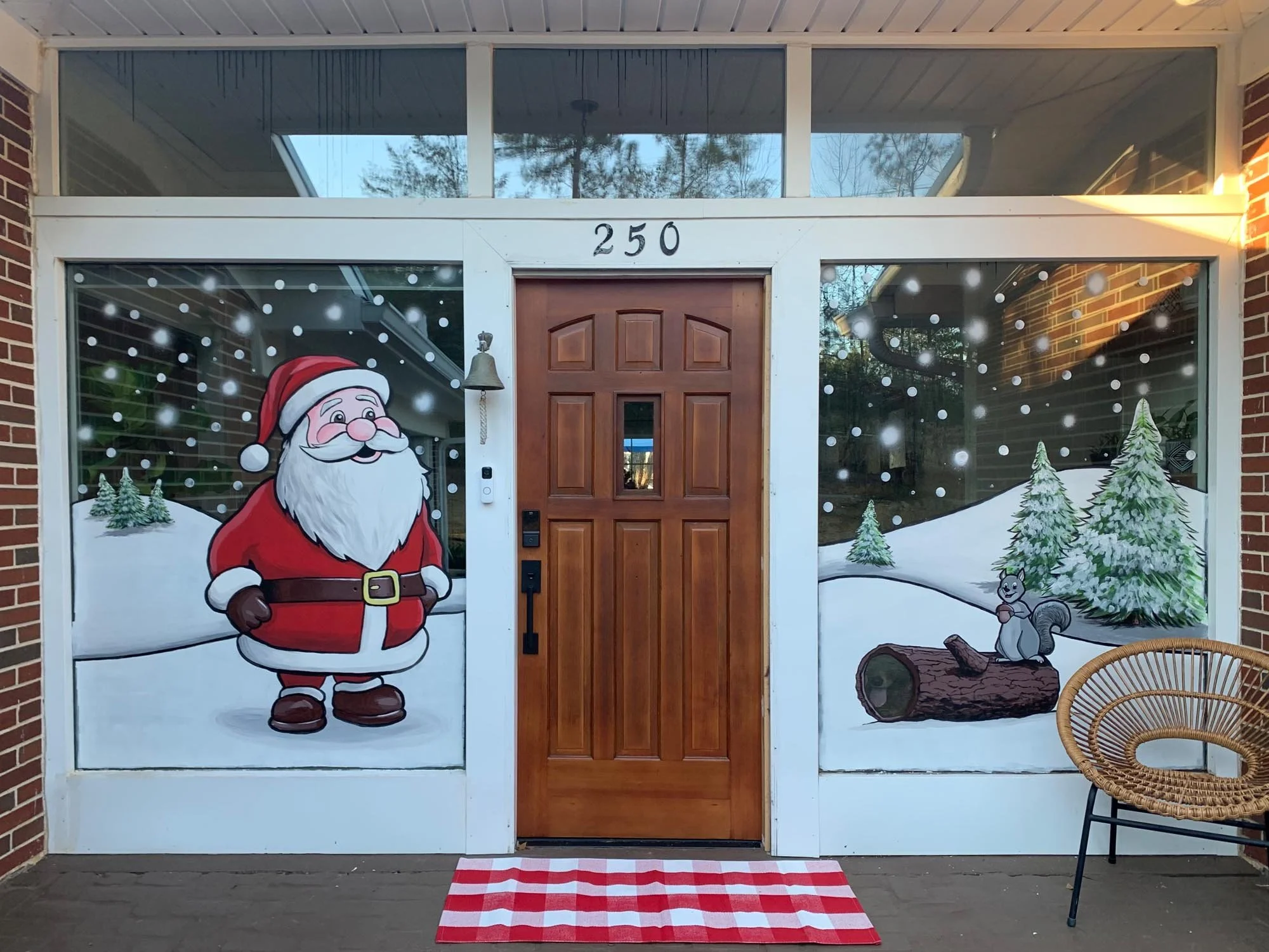 Decorative Christmas scene painted on window panels of front porch, featuring Santa Claus standing in a snowy landscape with pine trees, a squirrel on a log, snowflakes falling, and a red and white checkered rug in front of the door.