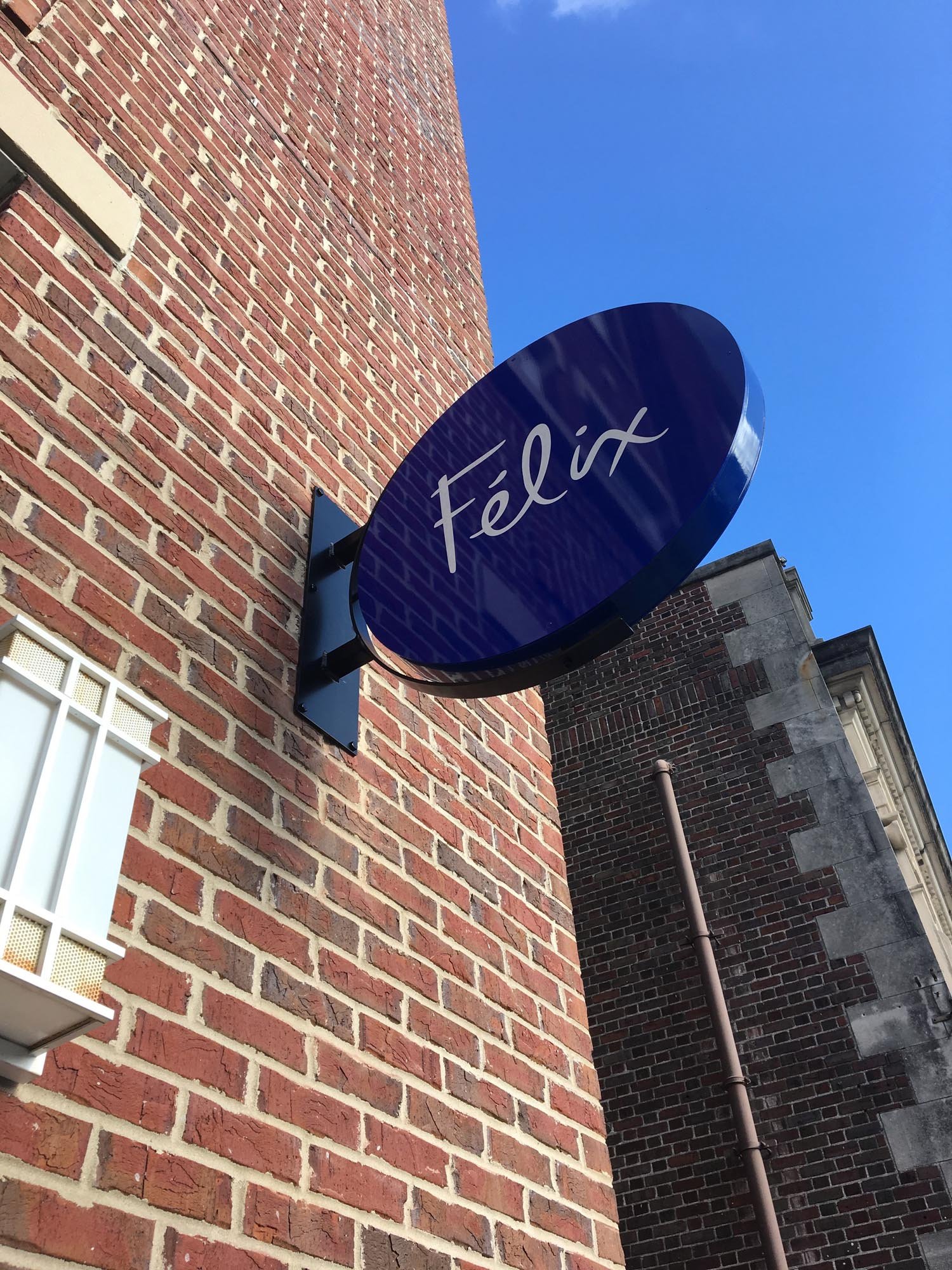 Blue oval sign with the word Félic in white cursive font mounted on a red brick building under a clear blue sky.