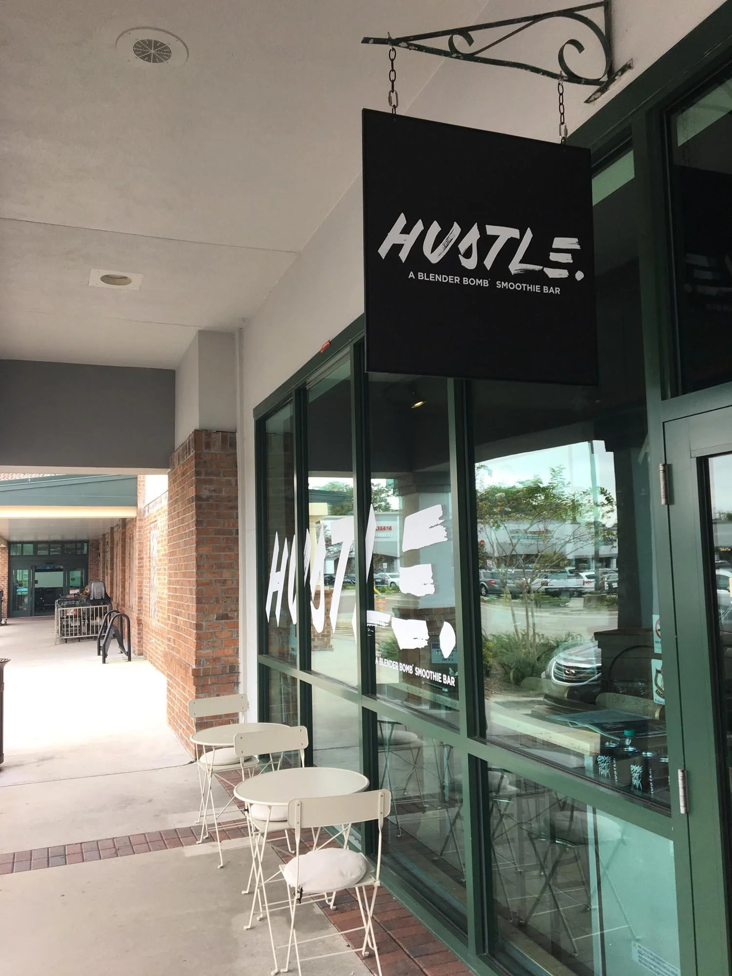 Exterior view of a smoothie bar called ``HUSTLE'' with outdoor seating, large windows, and a black sign hanging above the entrance.