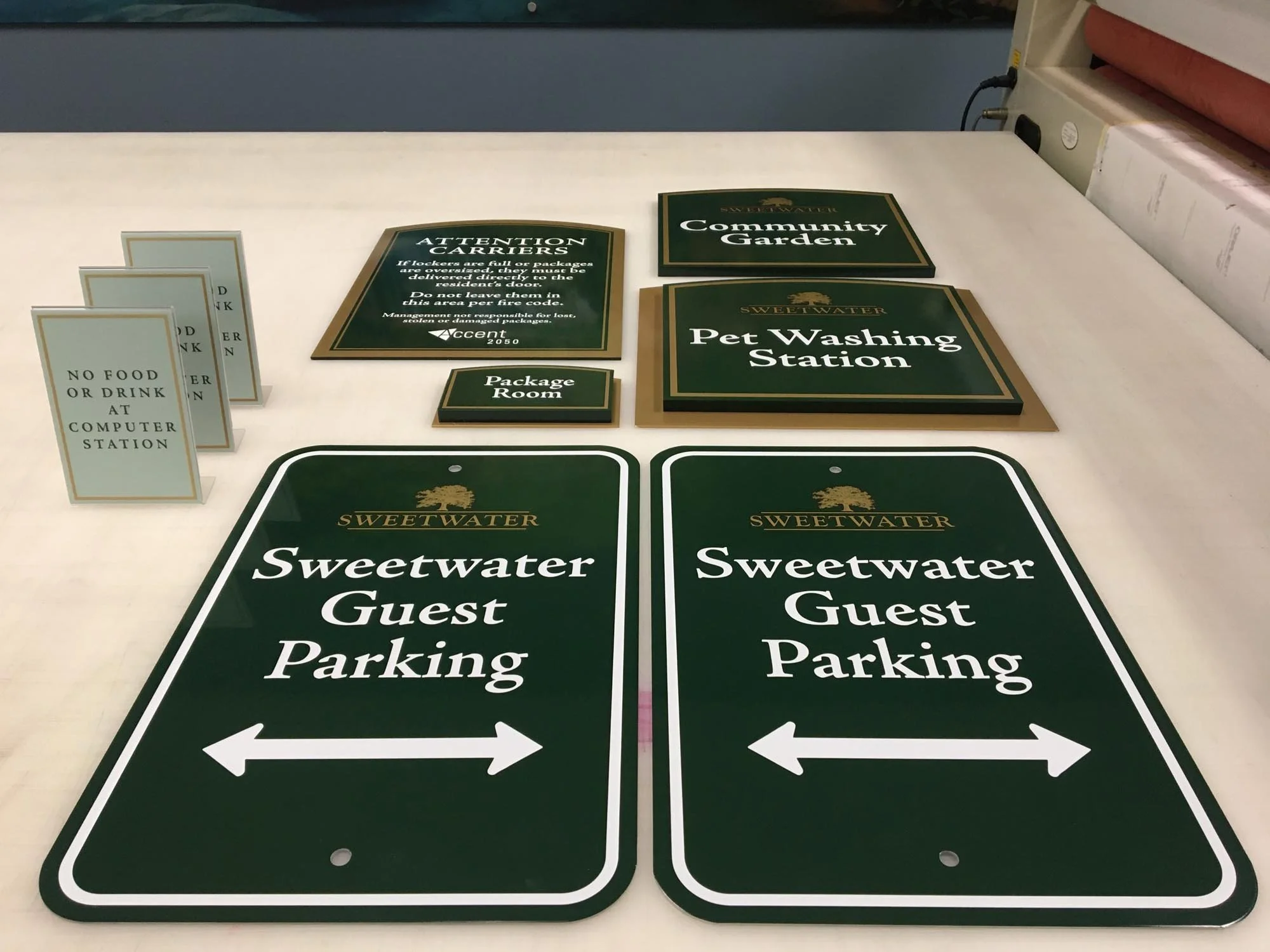 Green and white signs for Sweetwater guest parking, pet washing station, community garden, package room, and no food or drink at computer station.